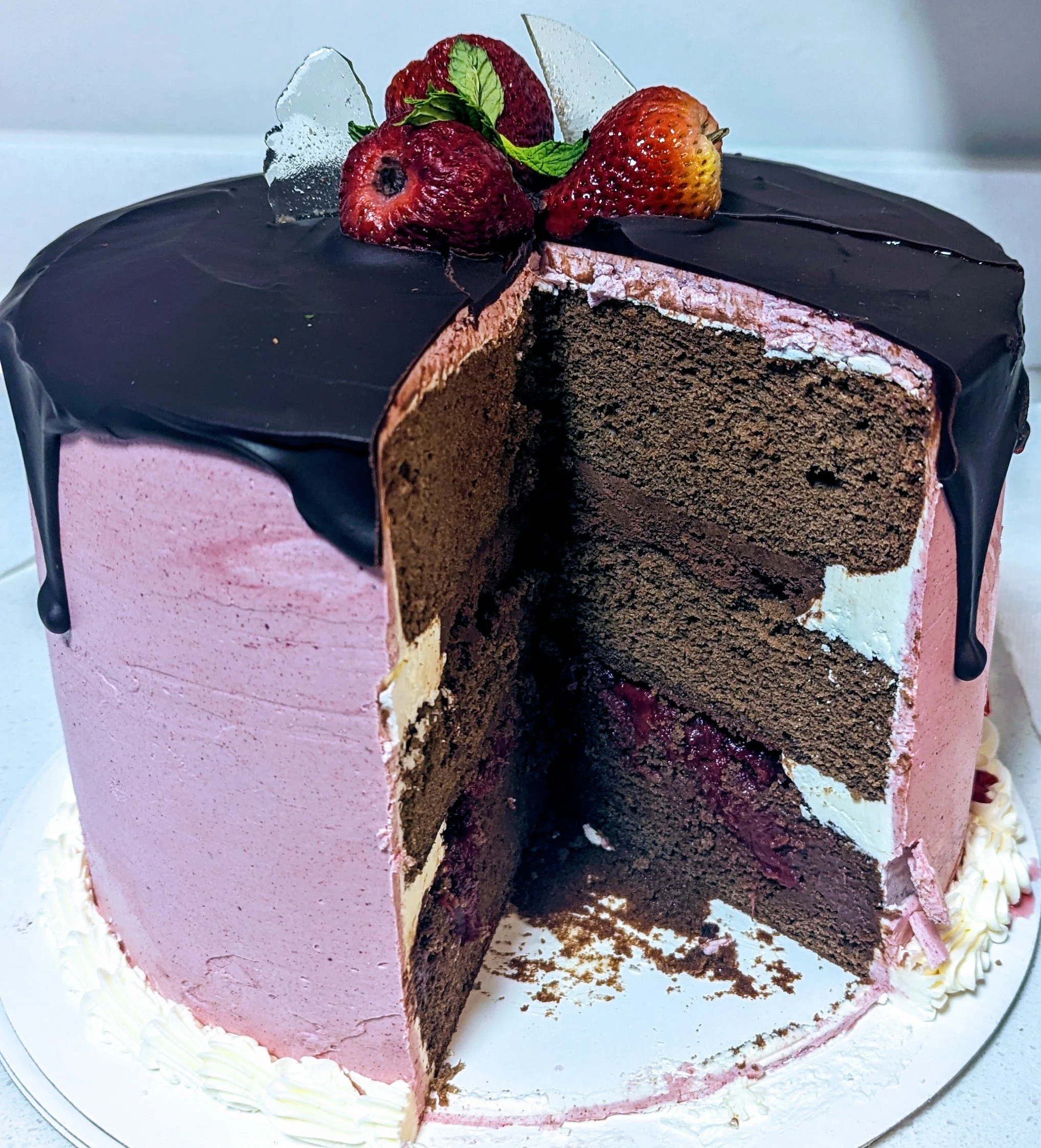 three tiered round chocolate chiffon cake with Italian buttercream pink frosting, glazed with chocolate ganache deluxe and topped with fresh strawberries, sprig of mint and sugar glass.