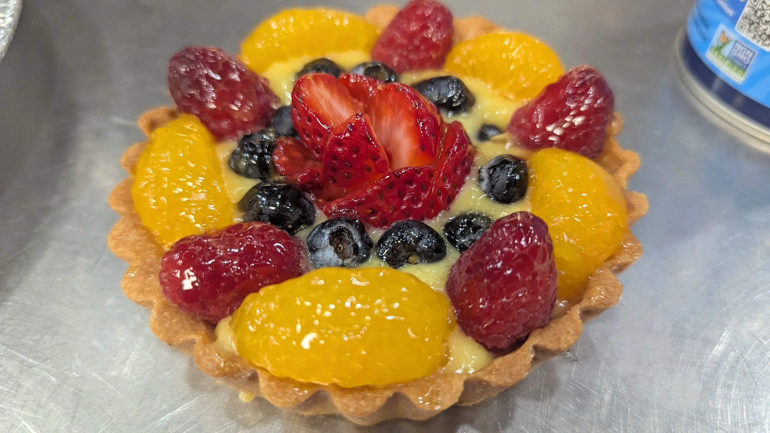 Fresh fruit tart with strawberries, blueberries, raspberries, and mandarins in a pastry crust on a metal surface.