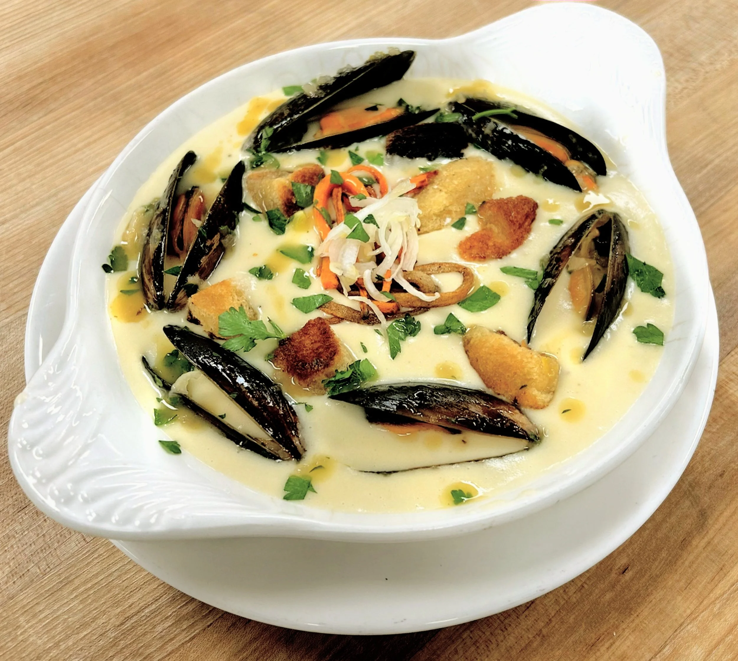 A bowl of creamy clam chowder garnished with chopped herbs, bread crumbs, and black mussels, served on a wooden table.
