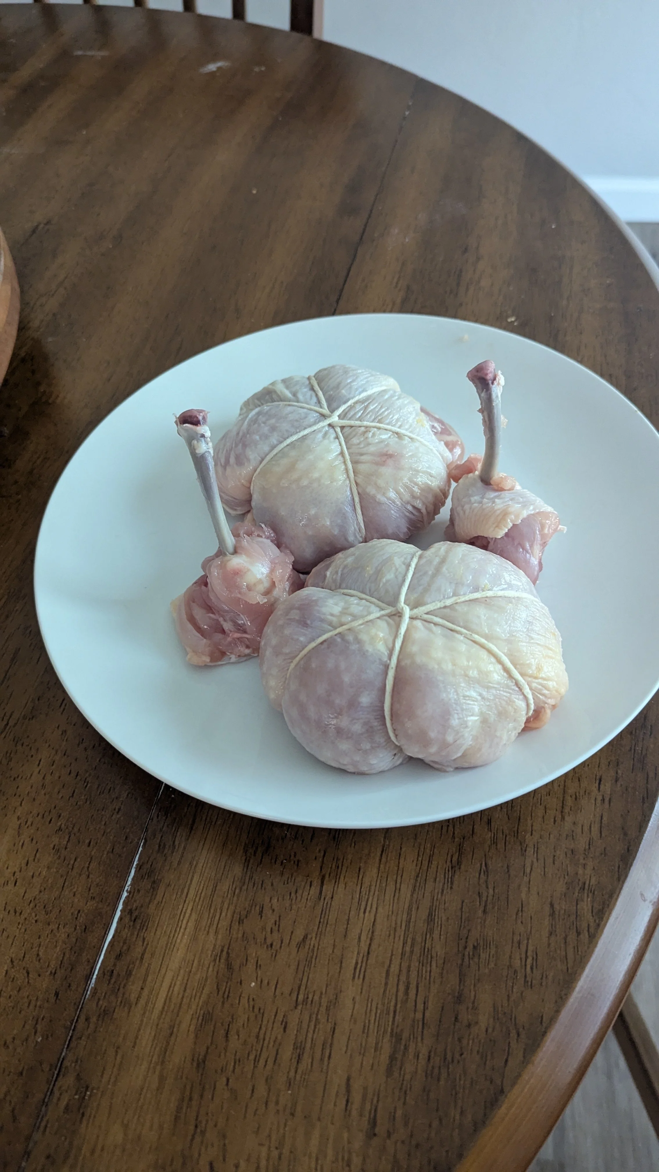 Two raw deboned chicken thighs tied with kitchen twine on a white plate on a wooden table, ready to roast, barbeque or broil.