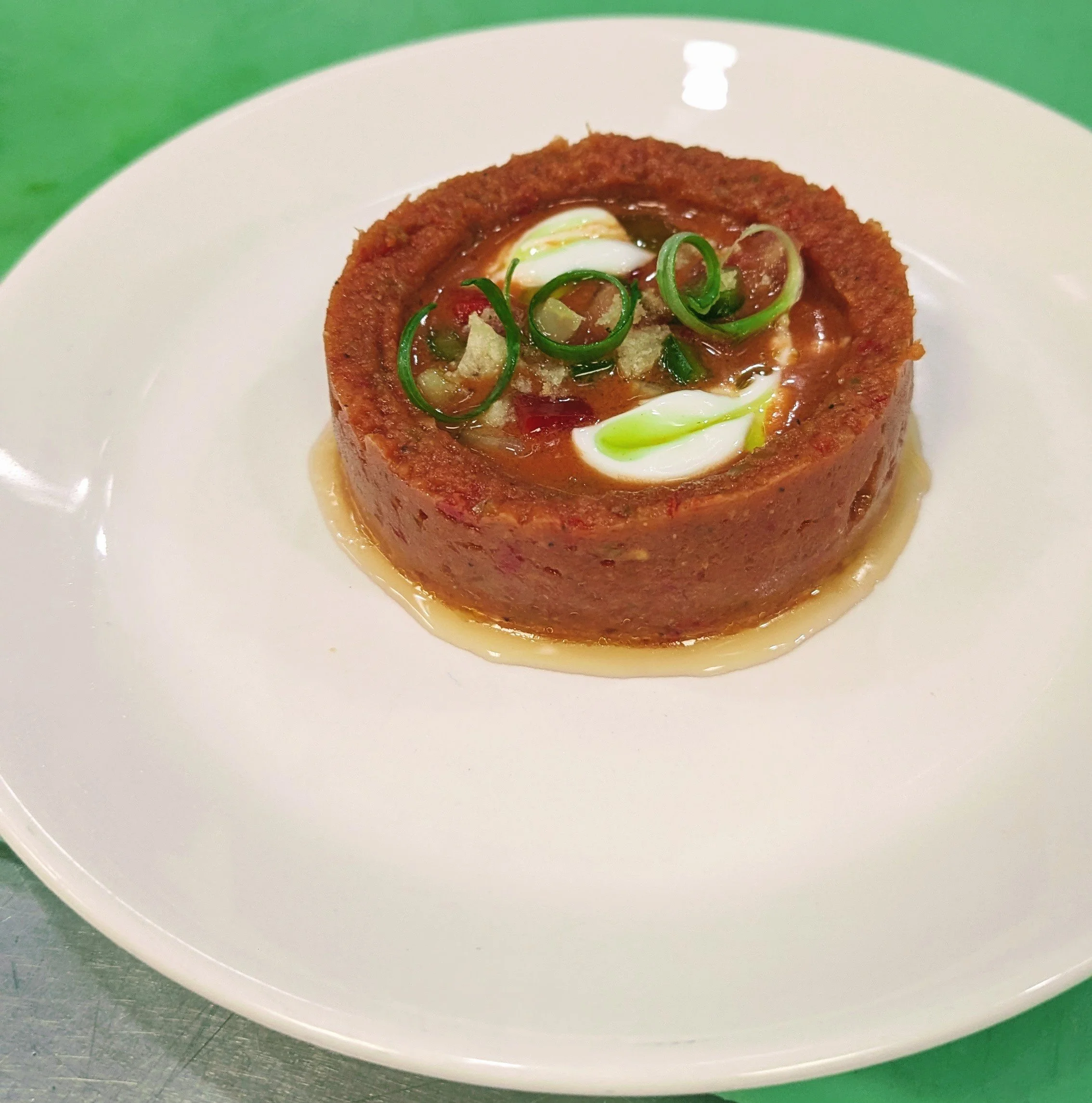 A round, fried tamale with a spicy filling, topped with green onion slices, chopped onions, and a drizzle of sauce, served on a white plate.