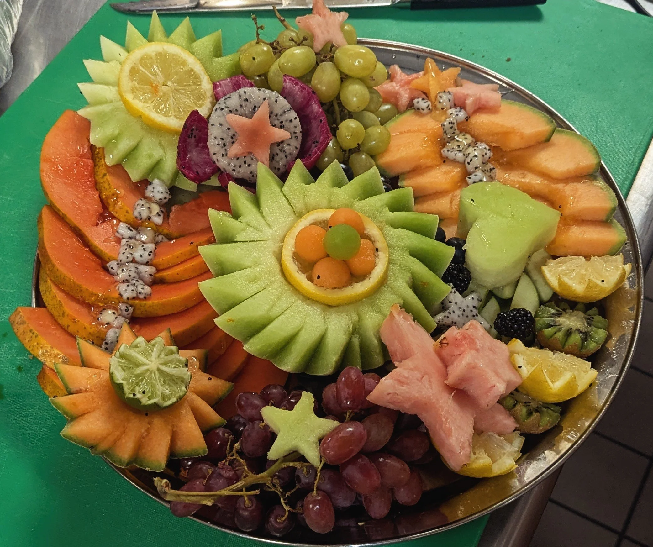 A large platter of assorted cut fresh fruits including melons, grapes, yellow and green citrus slices, kiwi, dragon fruit, star fruit, cantaloupe, honeydew, and pineapple, arranged in a colorful display.