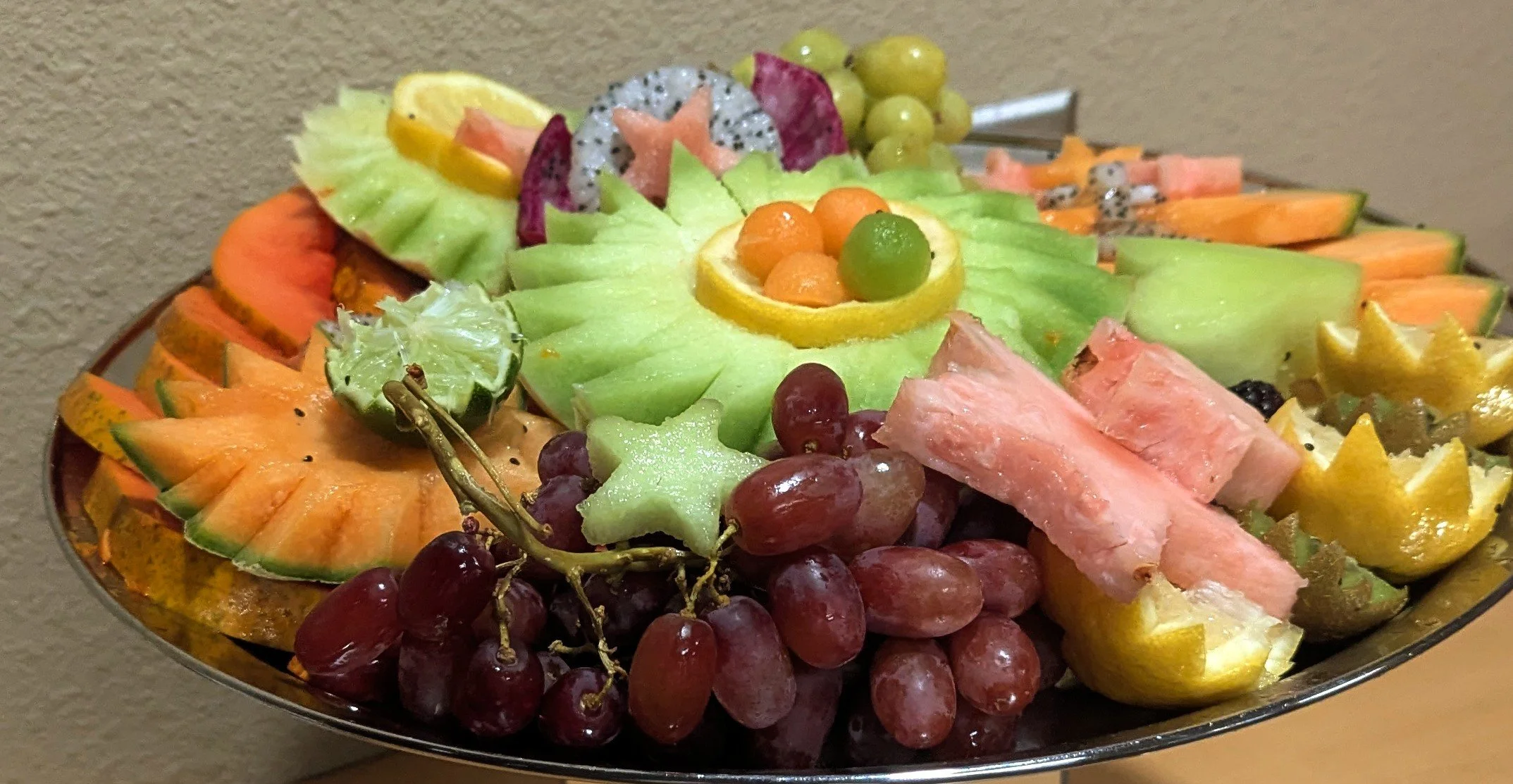 A silver platter filled with various sliced fresh fruits, including watermelon, cantaloupe, honeydew melon, grapes, dragon fruit, lemon slices, and star-shaped melon pieces.