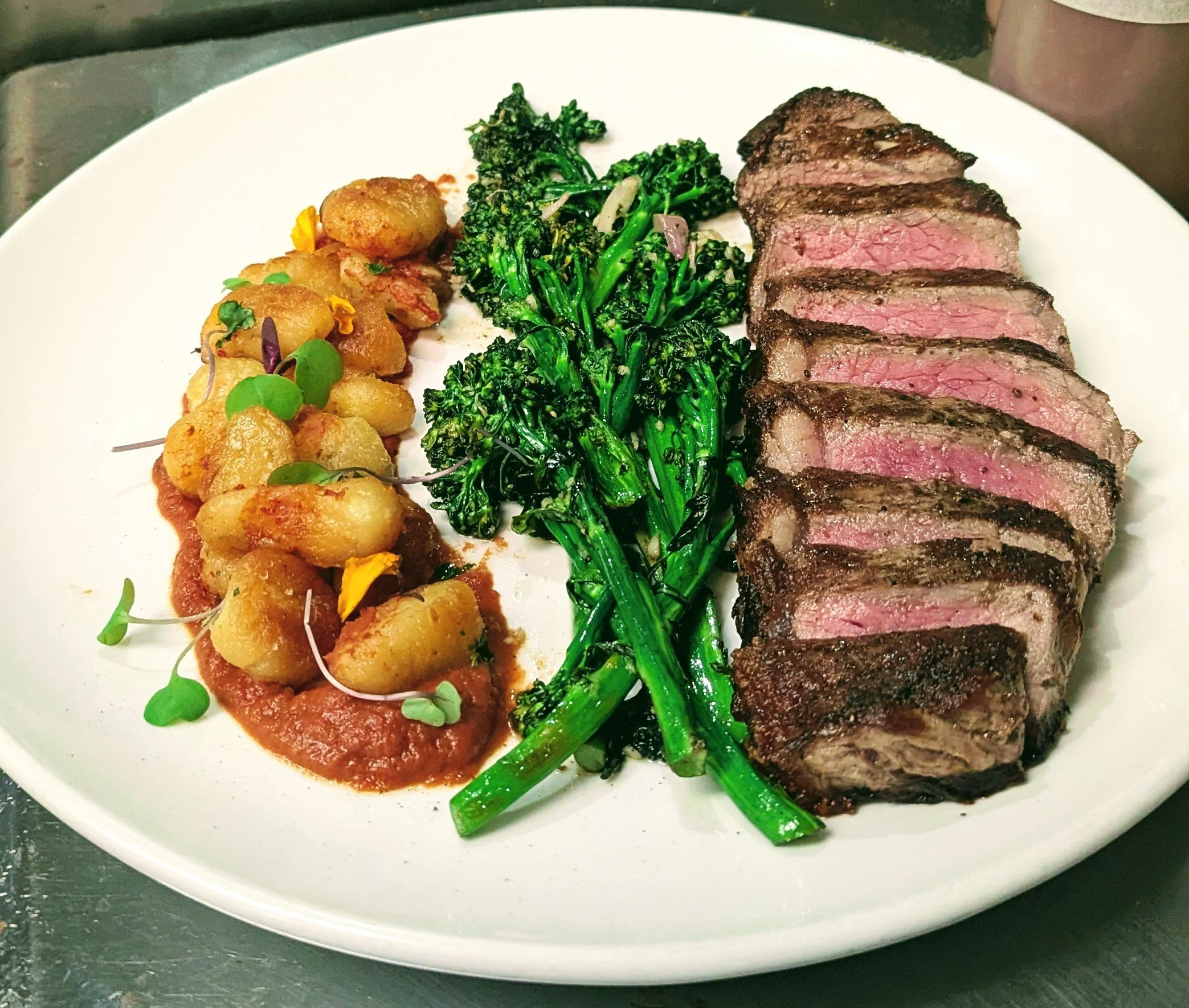 6 oz medium rare New York strip steak with crispy butter‑basted gnocchi, broccolini, and pistachio romesco, prepared by private chef Alexander Cole in Tucson, Arizona.