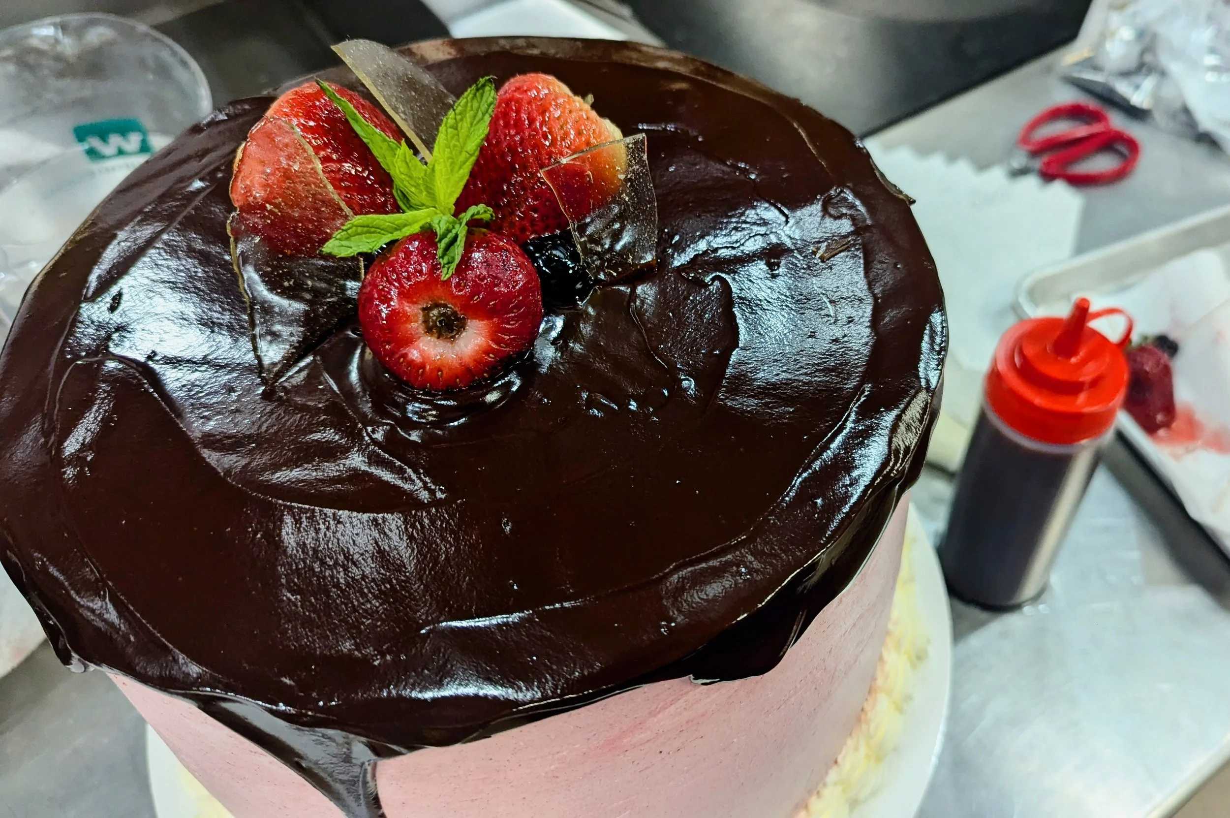 large round chocolate chiffon cake with Italian buttercream pink frosting, glazed with chocolate ganache deluxe and topped with fresh strawberries, sprig of mint and sugar glass.