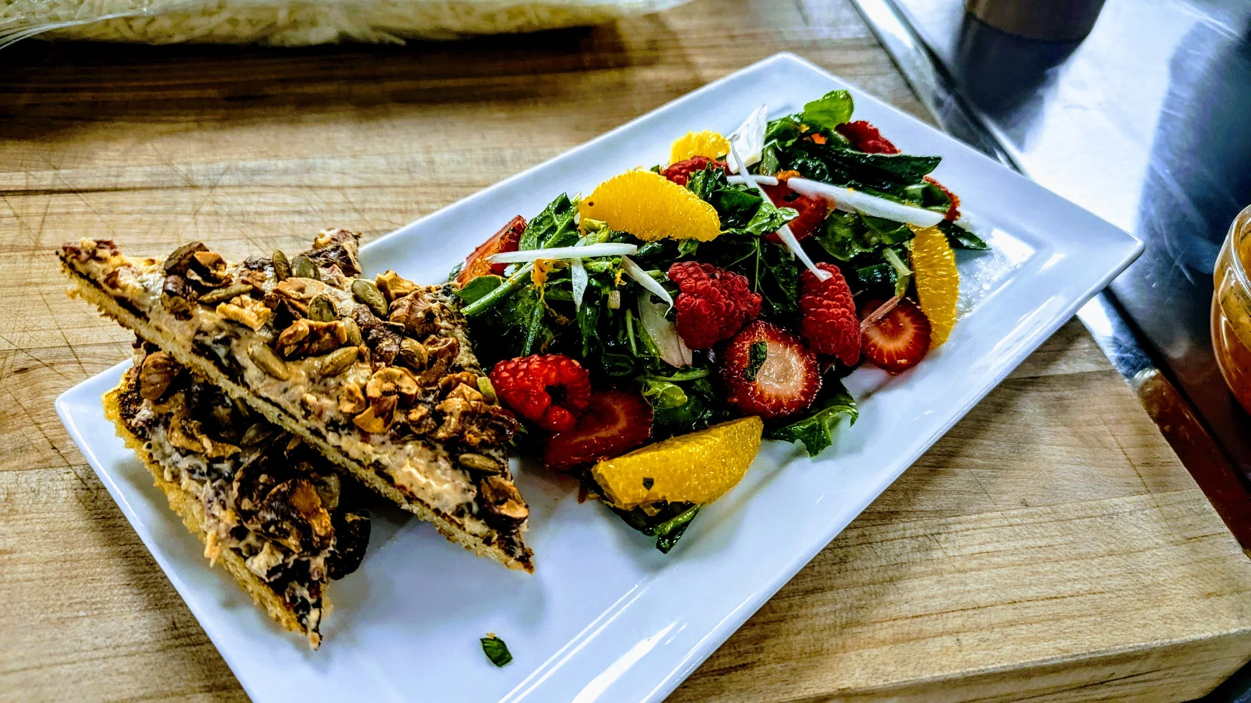 A white rectangular plate with two slices of layered dessert topped with chocolate and nuts on the left, and a fresh mixed berry salad with strawberries, raspberries, and orange slices on the right, on a wooden table.
