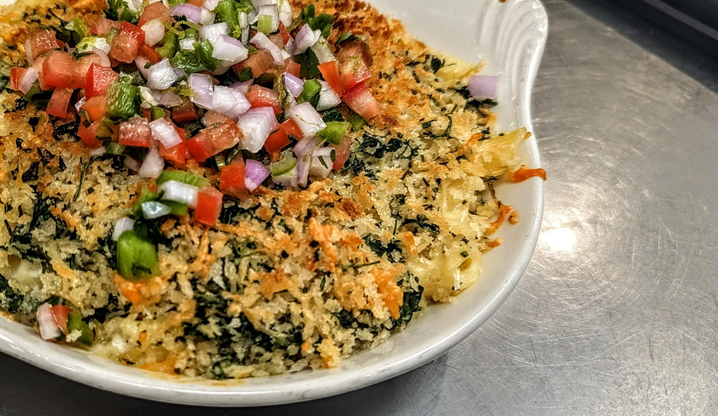 Baked casserole dish topped with diced tomatoes, onions, and green peppers, with a breadcrumb and herb crust, in a white oval baking dish on a metal surface.