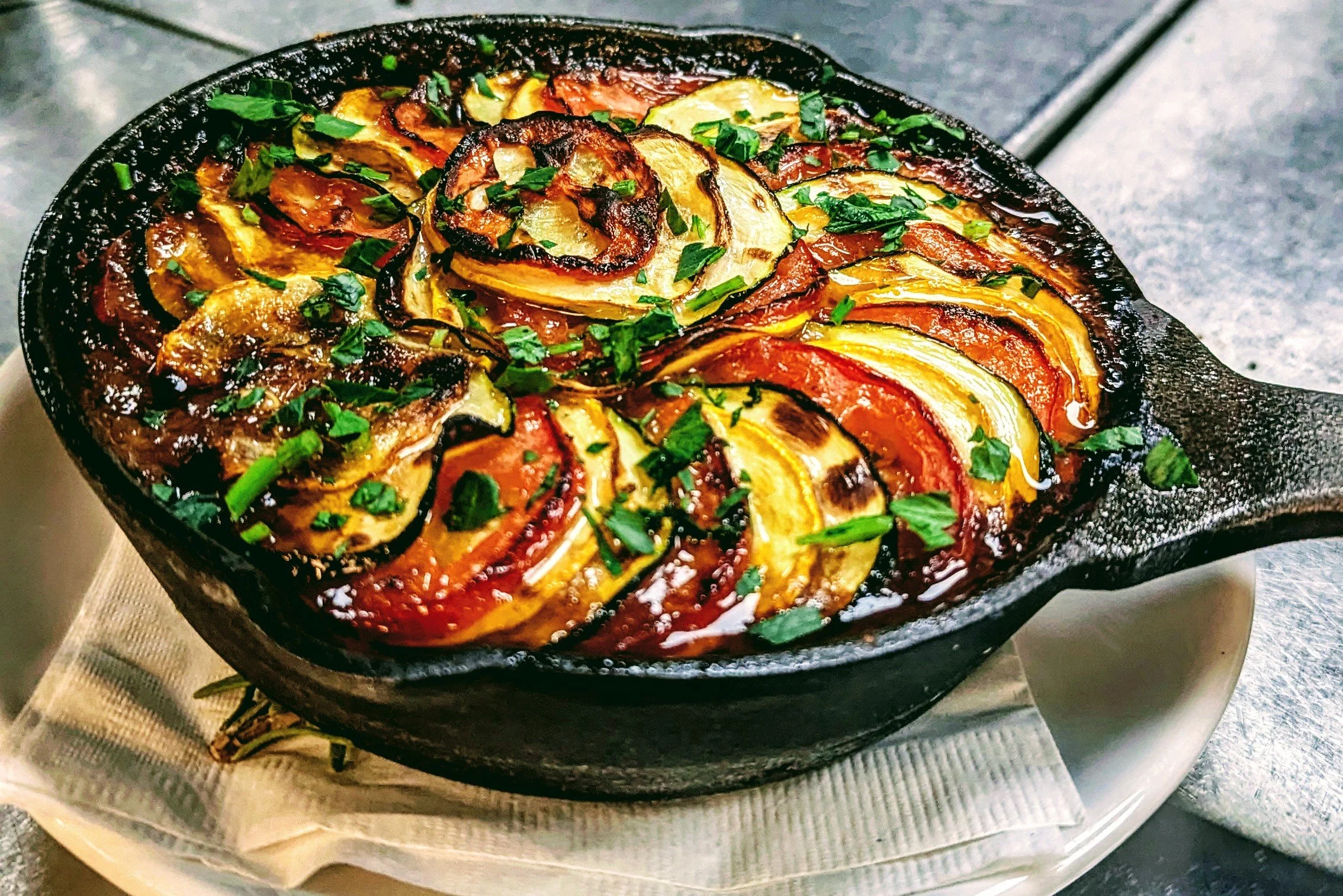 vegan ratatouille. Baked roasted vegetables in a cast iron skillet, including zucchini, yellow squash, and tomatoes, tomato sauce pomodoro, drizzled extra virgin olive oil, garnished with parsley, chives and sage.