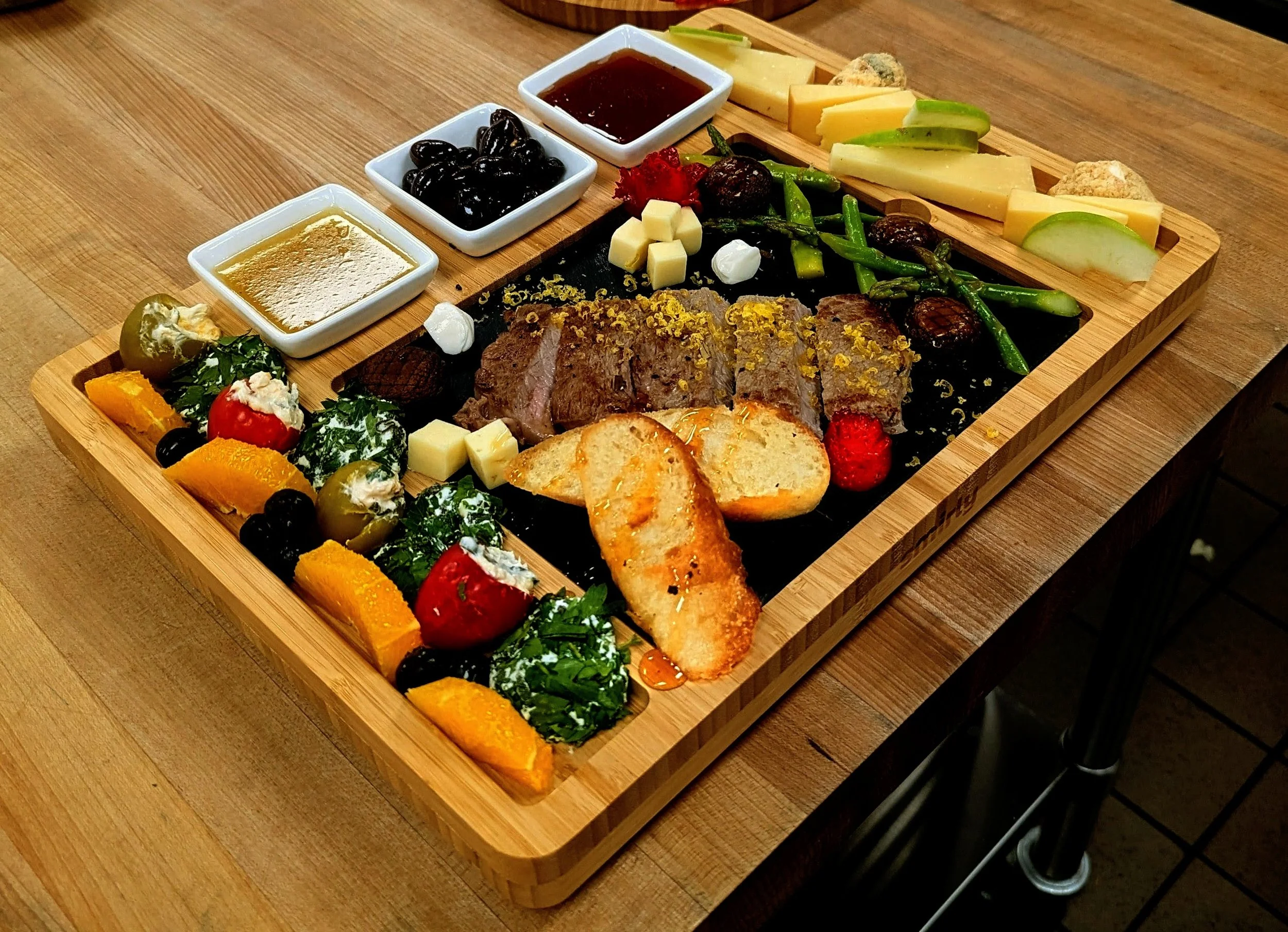A large wooden board with a variety of food items including sliced beef, vegetables, cheeses, olives, sauces, bread, and garnishes arranged for serving.
