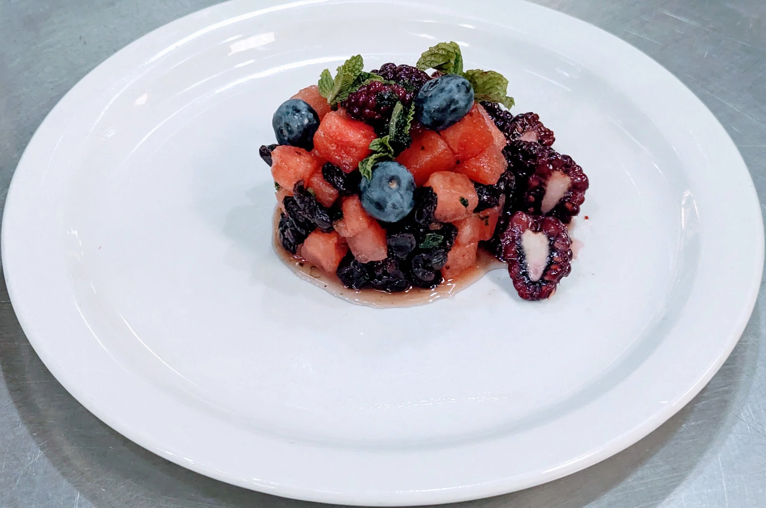 A fruit salad with watermelon, blueberries, blackberries, and mint leaves on a white plate.