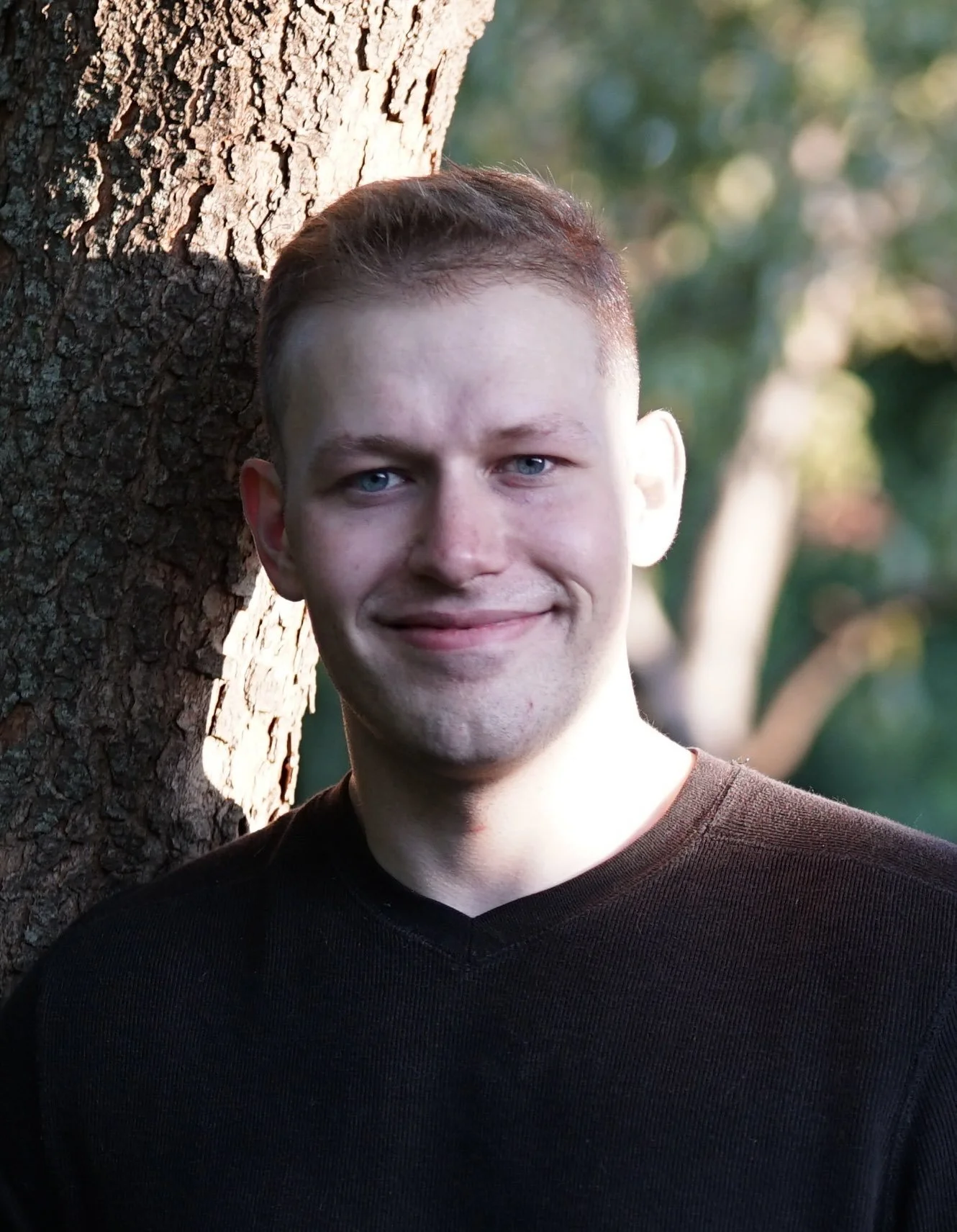 A young man with light skin, blue eyes, and short brown hair, smiling, standing outdoors next to a tree with sunlight on his face.
