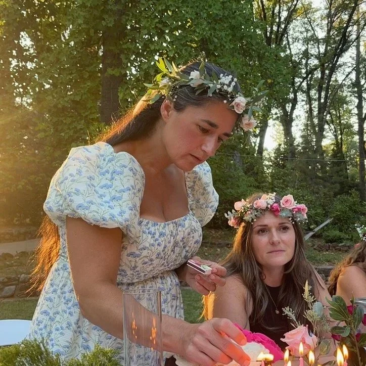 Two women wearing floral crowns at an outdoor gathering during sunset, with one lighting candles.