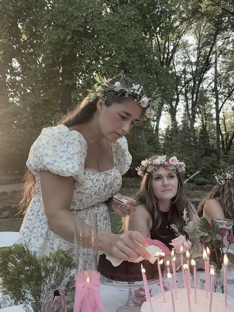 Women with flower crowns celebrating at a table with a pink birthday cake and lit candles outdoors.