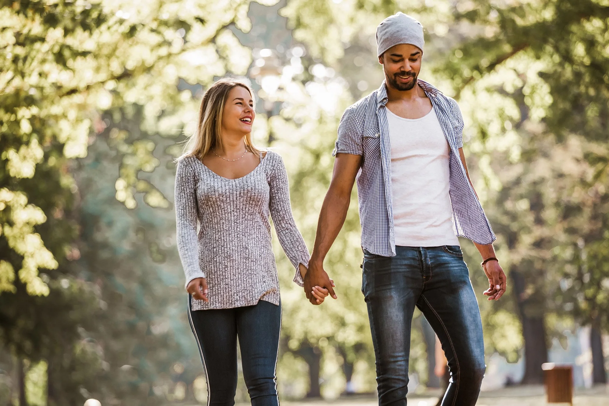A couple holding hands, walking through a park with trees and sunlight in the background, smiling and enjoying each other's company.