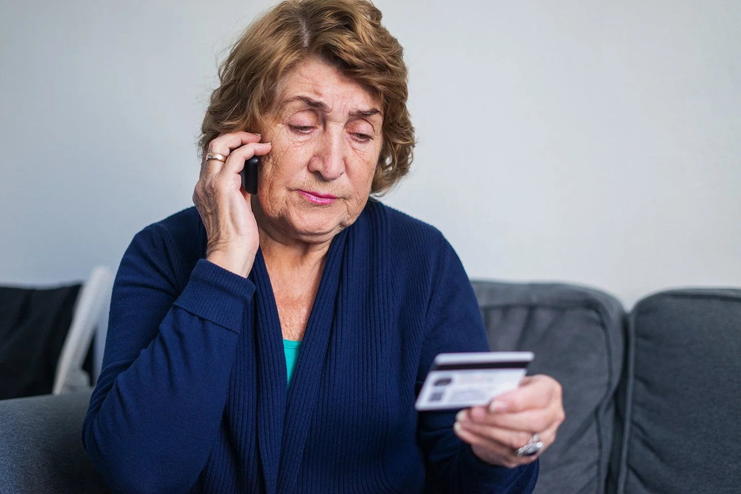 Older woman sitting on a gray couch, talking on a mobile phone with her left hand and holding a credit card in her right hand, wearing a dark blue cardigan.