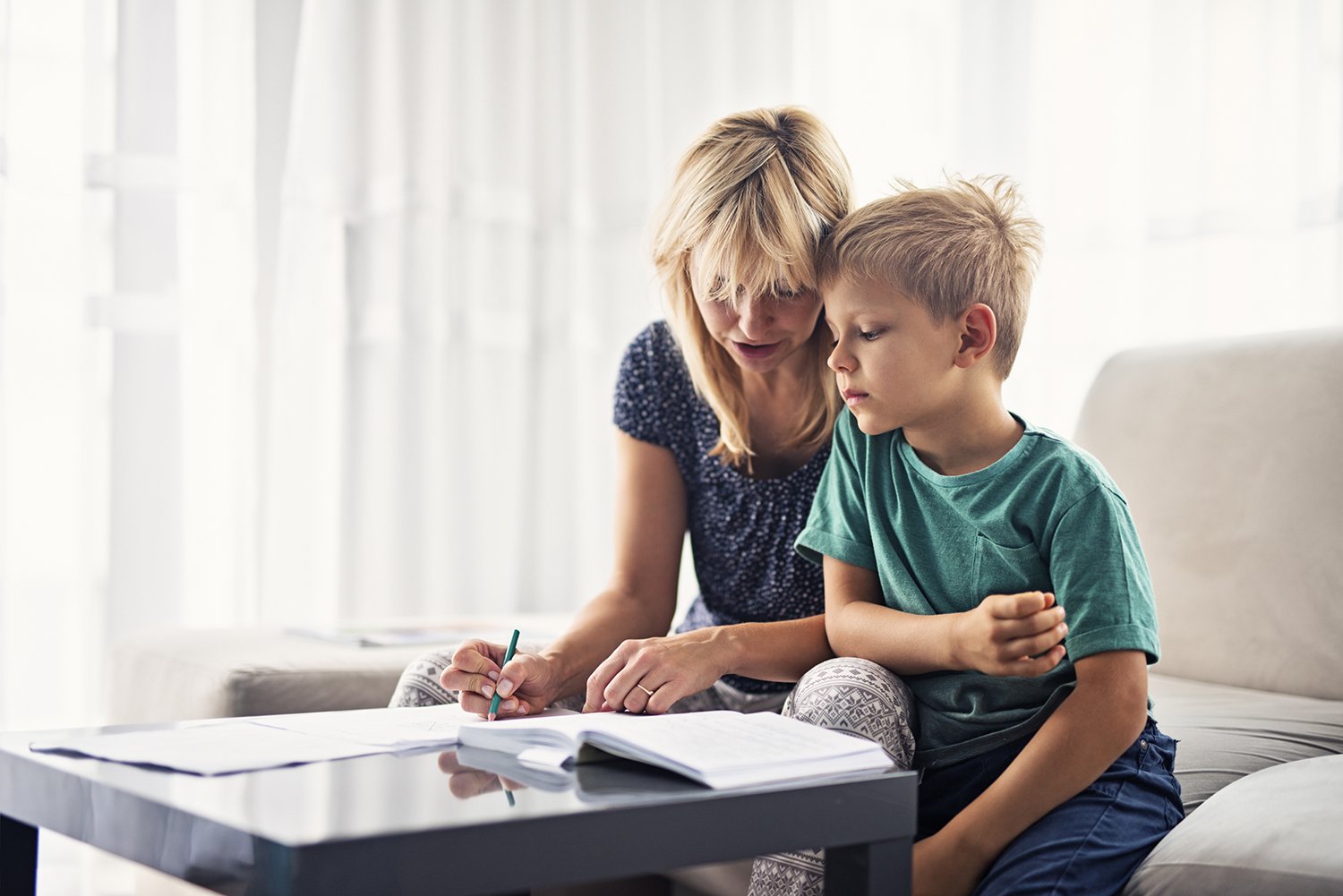 A woman and young boy sitting together on a couch in a bright room, looking at an open book on a table, with the woman pointing at the book and the boy looking focused.