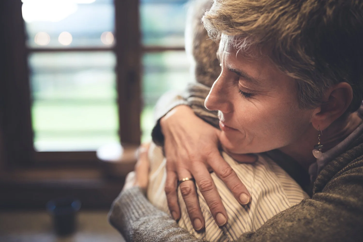 A woman hugging a man with her eyes closed, showing emotional comfort, indoors with sunlight coming through window blinds.
