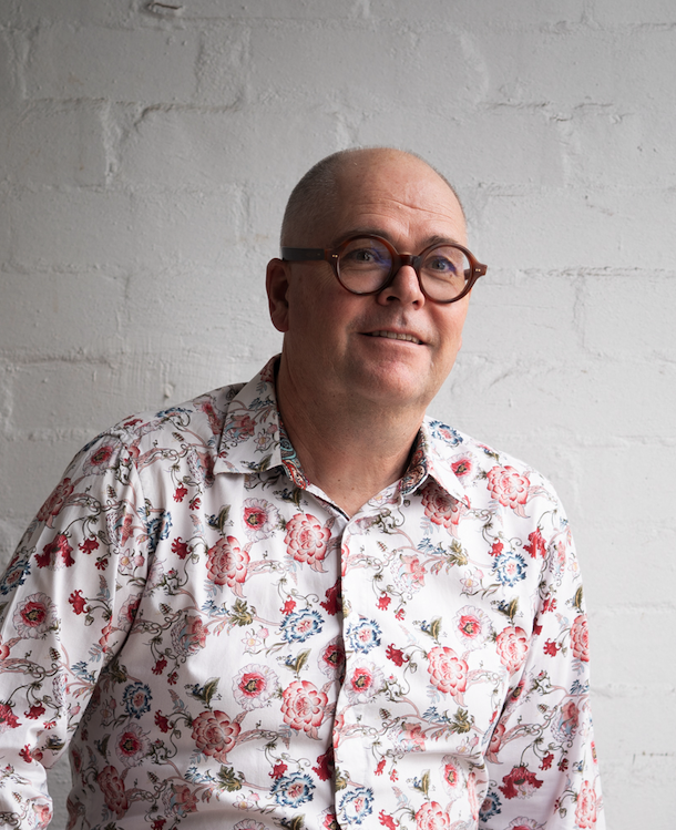A middle-aged man with glasses and a floral shirt standing against a white brick wall.