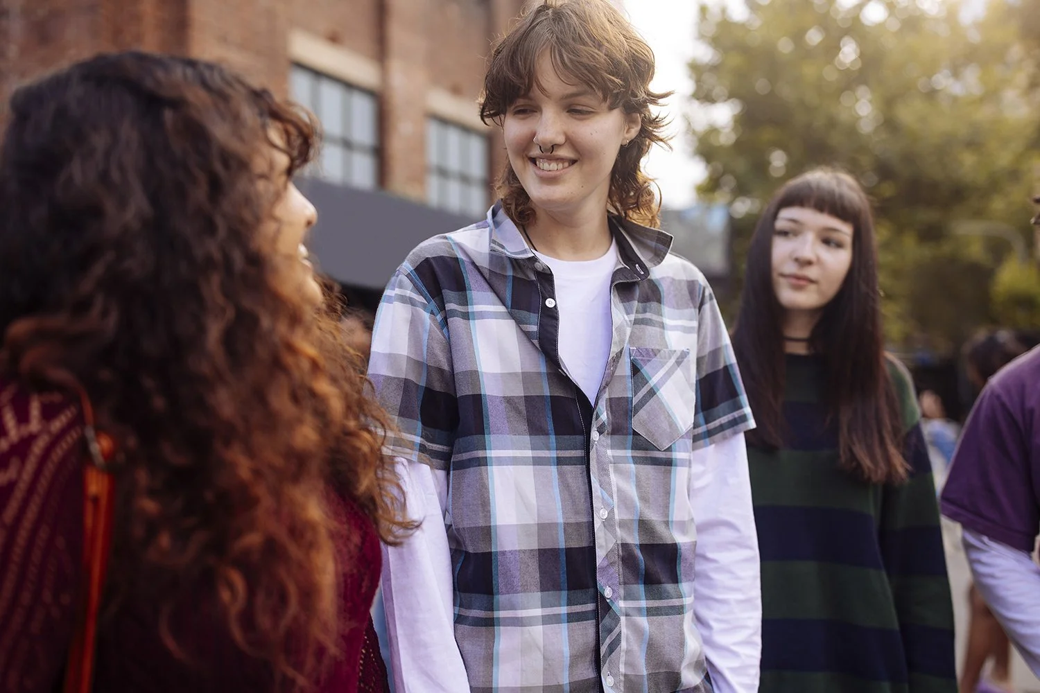 Four young women talking outdoors, with trees and a brick building in the background.