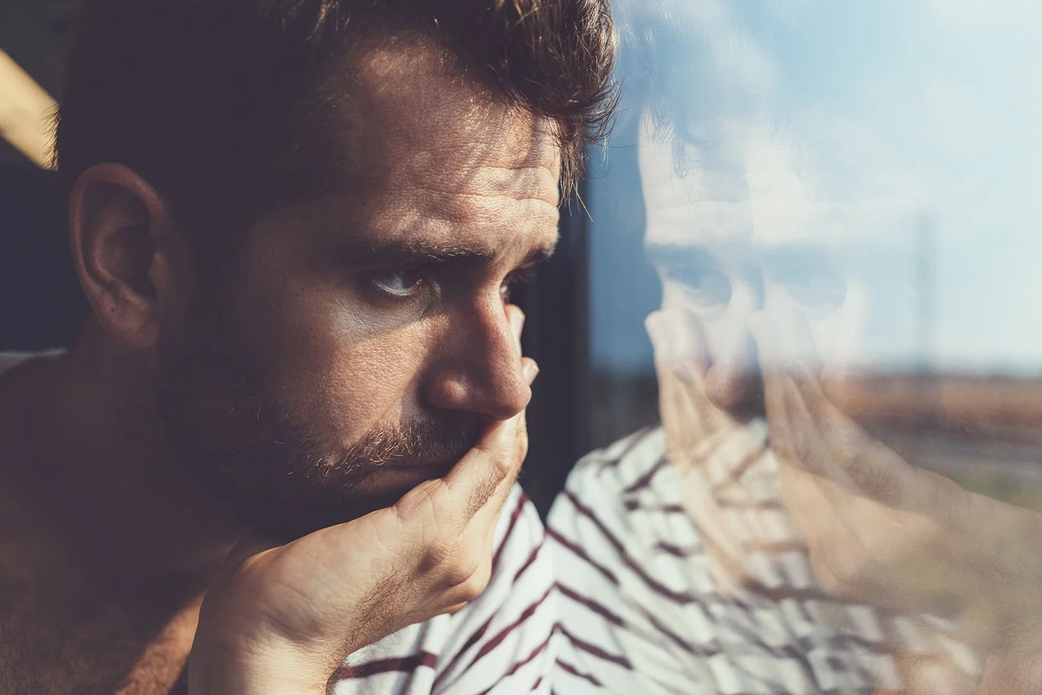 A man with short brown hair and a beard looks thoughtfully out the window, resting his face on his hand, with his reflection visible on the glass.