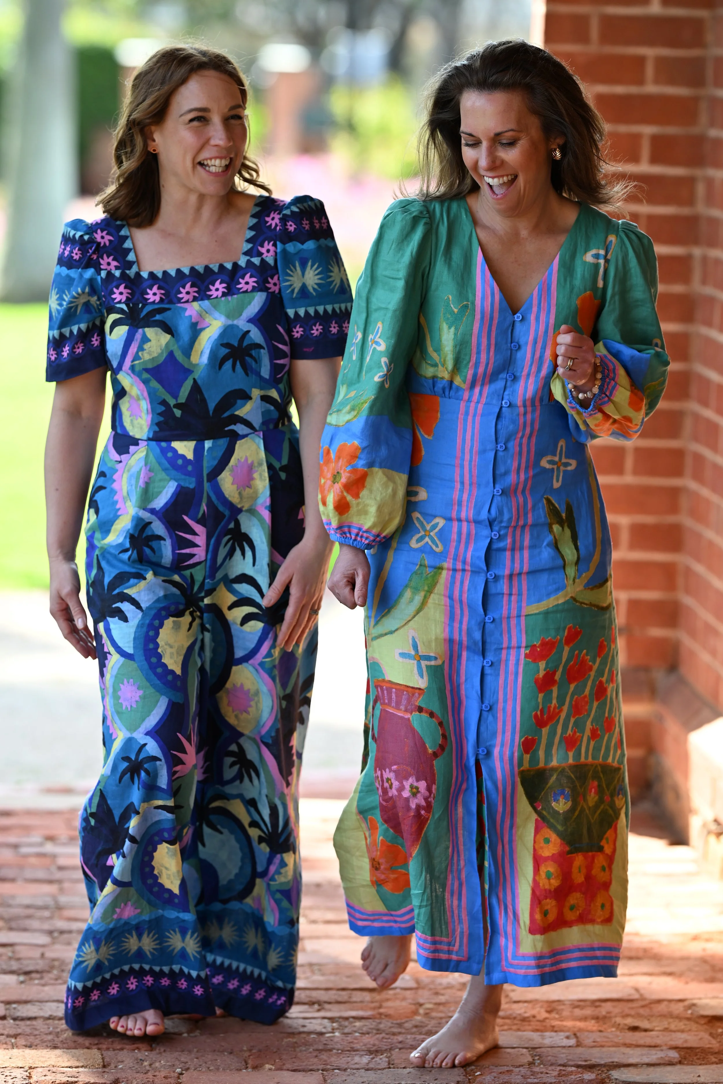 Two women walking outdoors, smiling and laughing, wearing colorful, patterned maxi dresses. One woman has short brown hair and the other has long, gray hair. They are barefoot on a brick path, with a brick wall and greenery in the background.