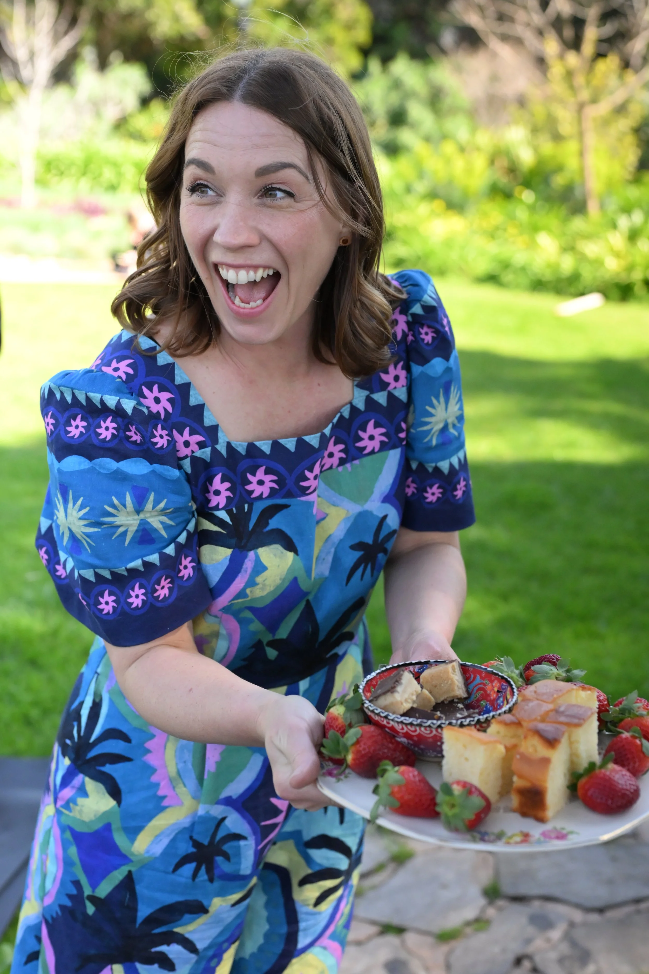 A woman in a colorful blue dress with floral patterns is smiling and holding a plate of desserts and strawberries outdoors in a garden.