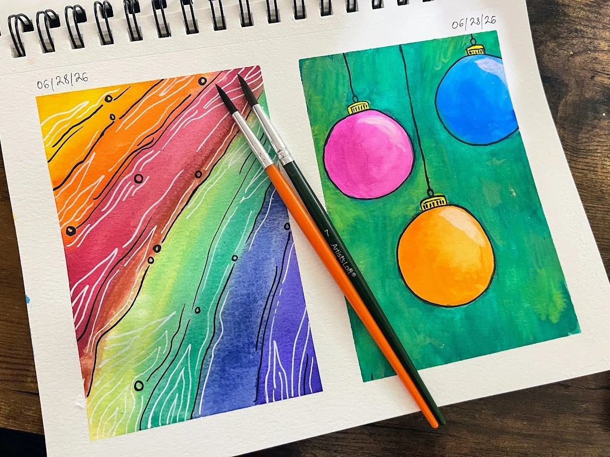 Colorful watercolor drawings on a sketchbook with a rainbow pattern on the left and hanging Christmas ornaments on the right, dated 06/28/26, with two paintbrushes resting on the pages.