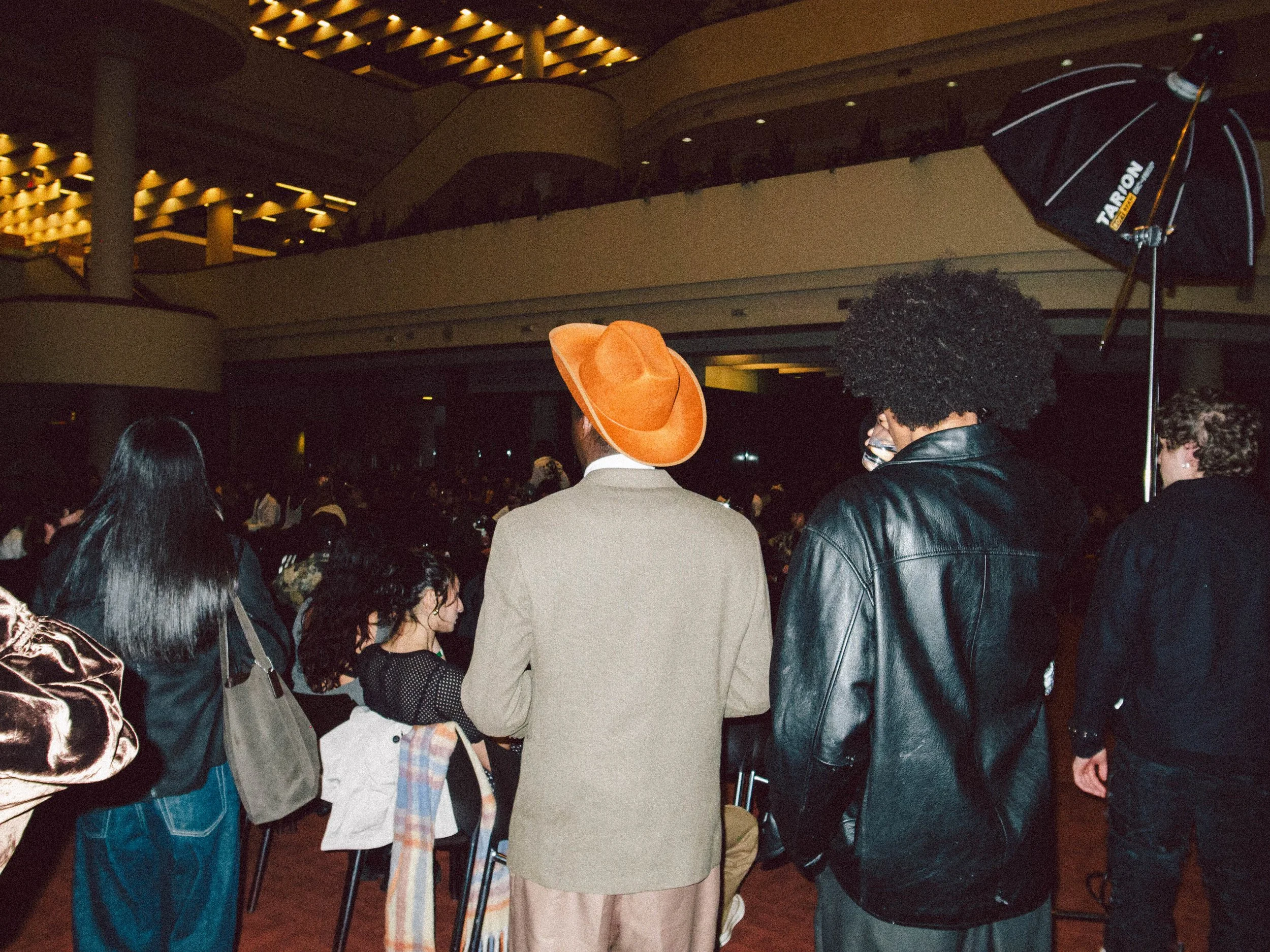 People attending an indoor event, some wearing stylish clothing and hats, with a focus on three individuals in the foreground, one with an orange hat, another with curly hair and a leather jacket, and a third with a wig or hairstyle, in a dimly lit v