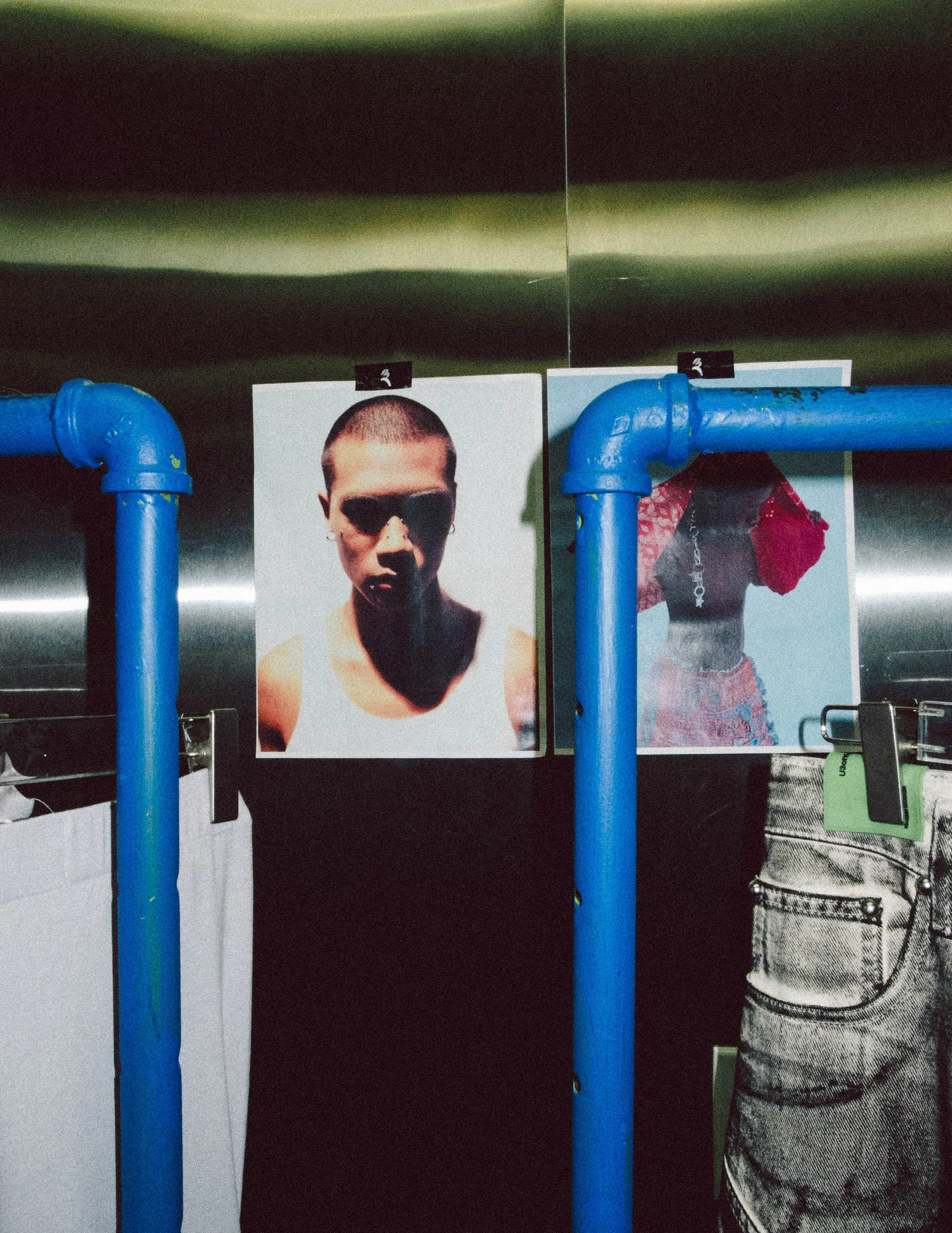 Photographs of a person with a shaved head and a person with a red hat are hanging on a metallic wall, separated by blue pipes.