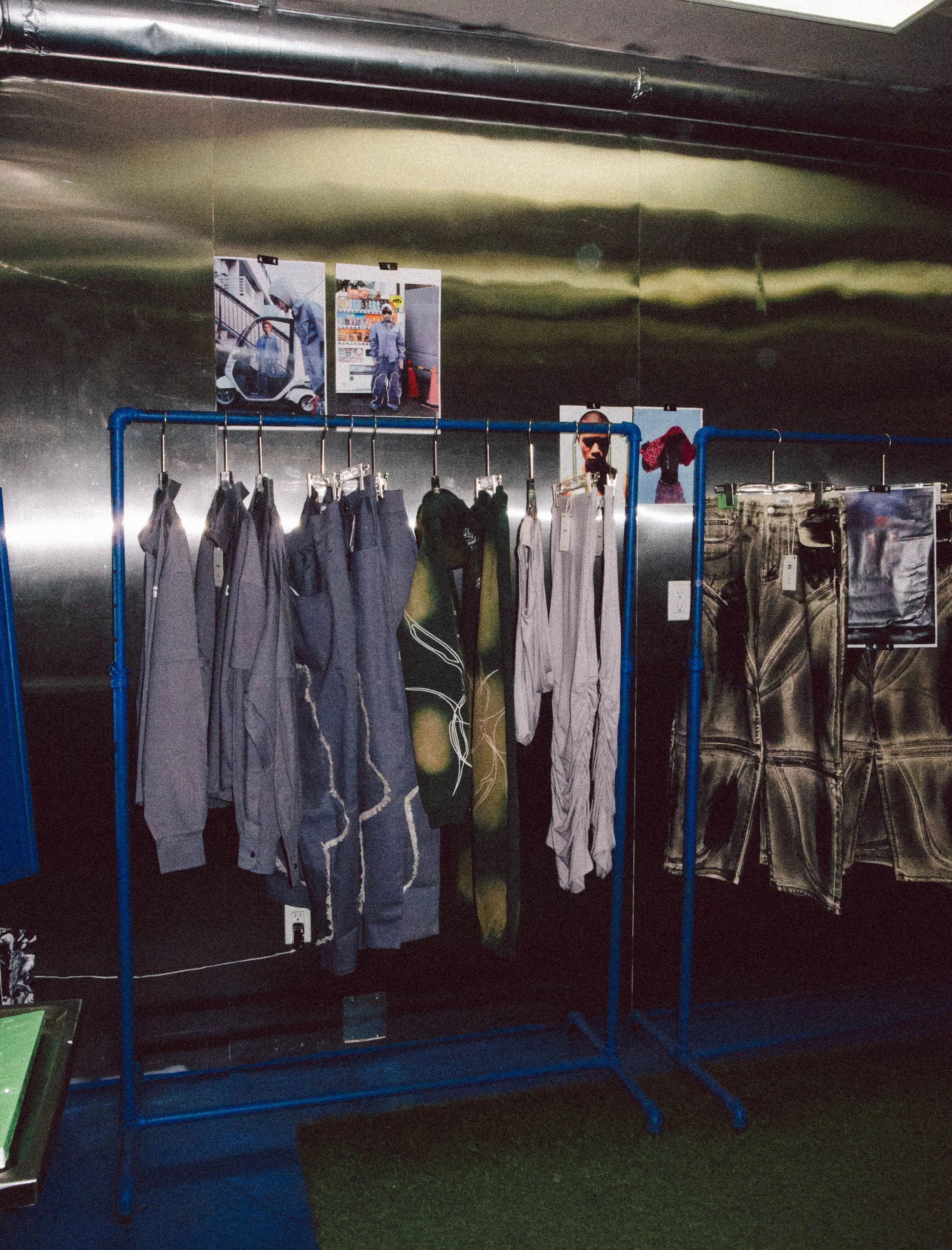 Clothing rack displaying jackets, hoodies, and pants in an indoor space with posters on the wall above.