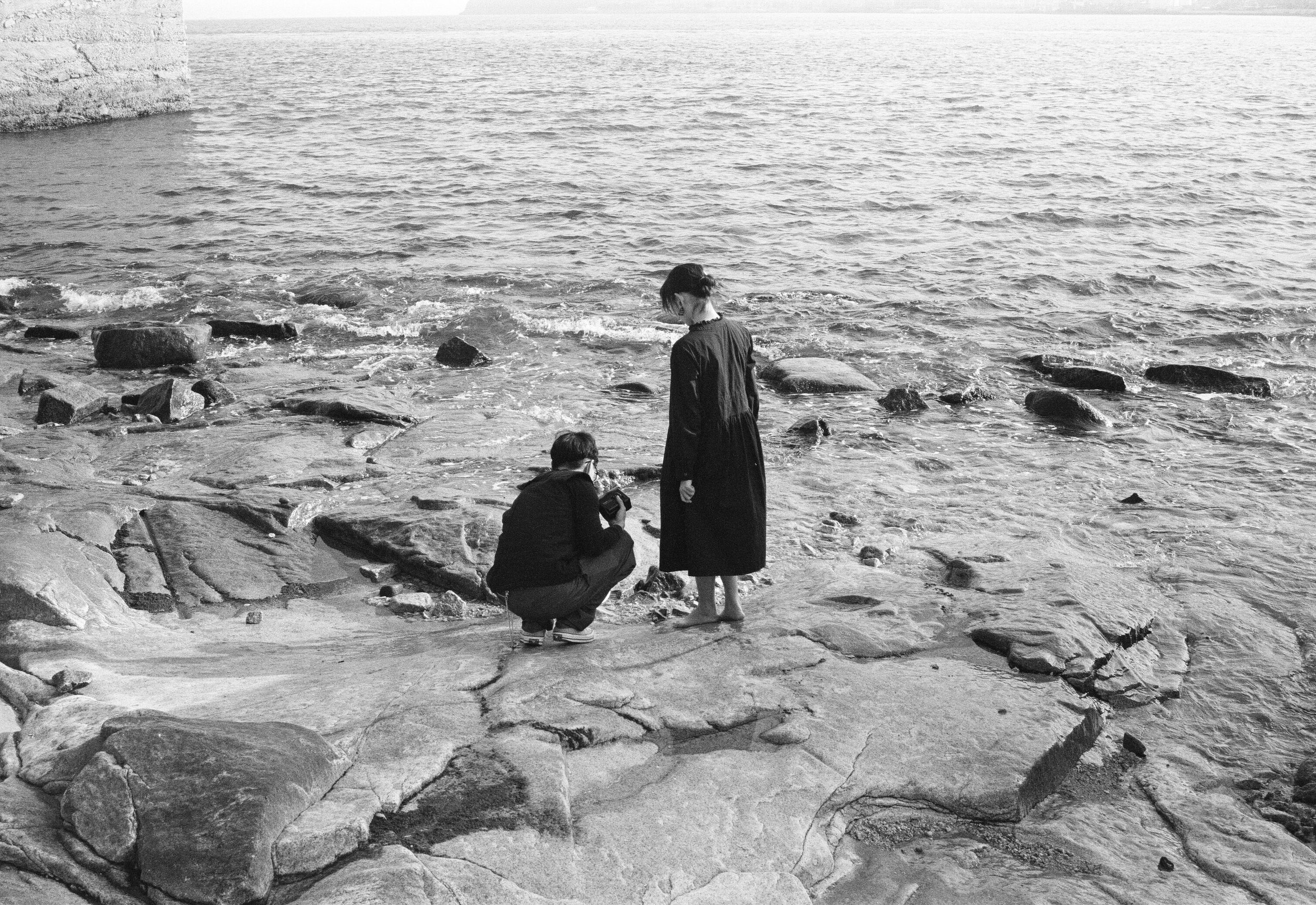 a lovley couples near the shoreline, one standing and one crouching, looking at the water, with rocks and the sea in the background.