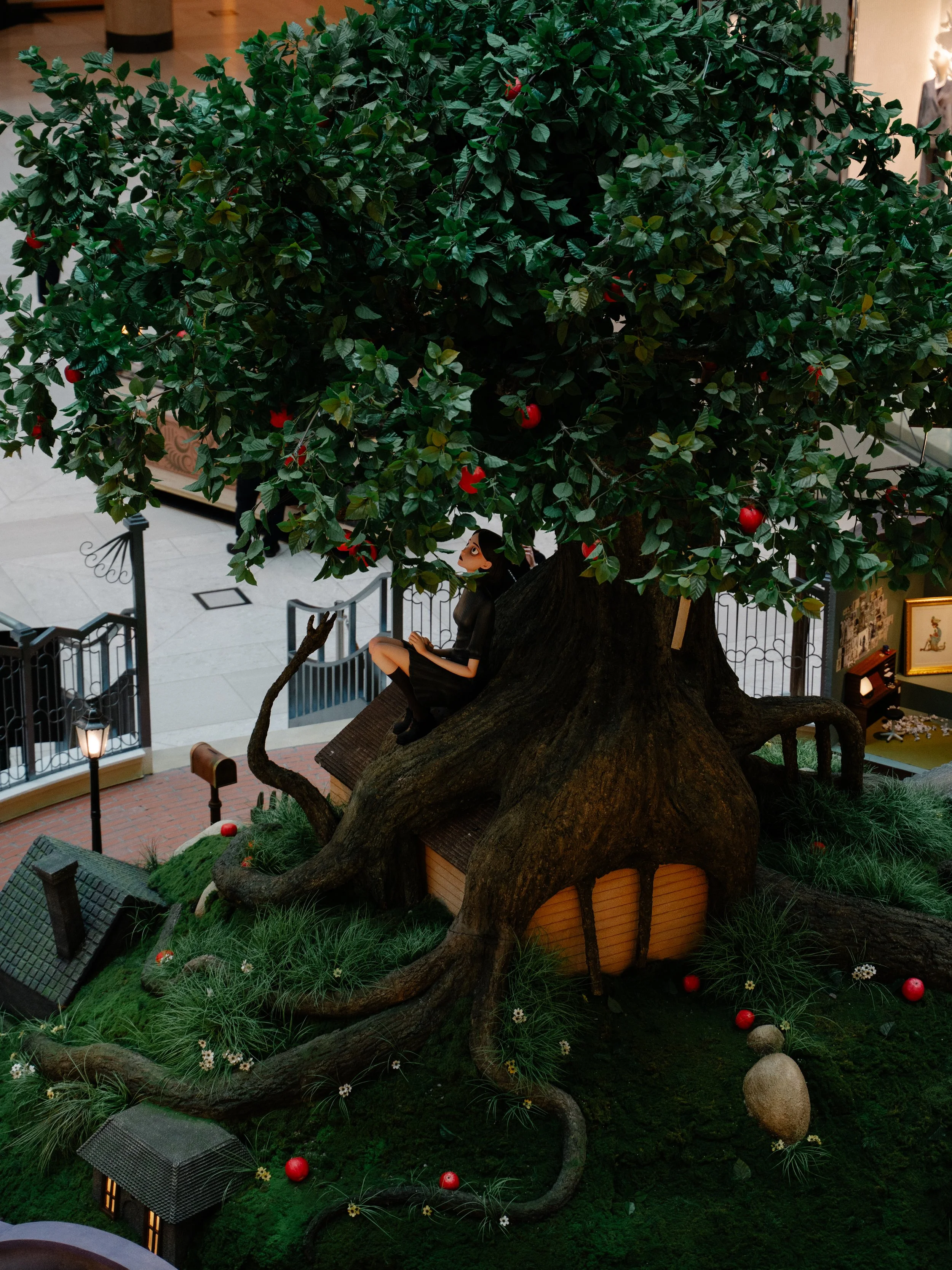 A large decorative tree with green leaves and red apples, with a small house and a woman in black sitting on a ledge under the tree, displayed indoors.
