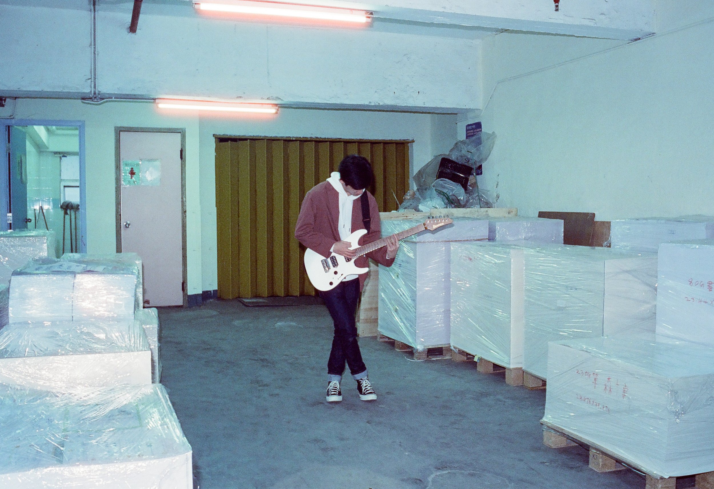 A person playing a white electric guitar in an industrial storage area with wrapped pallets and a yellow door.