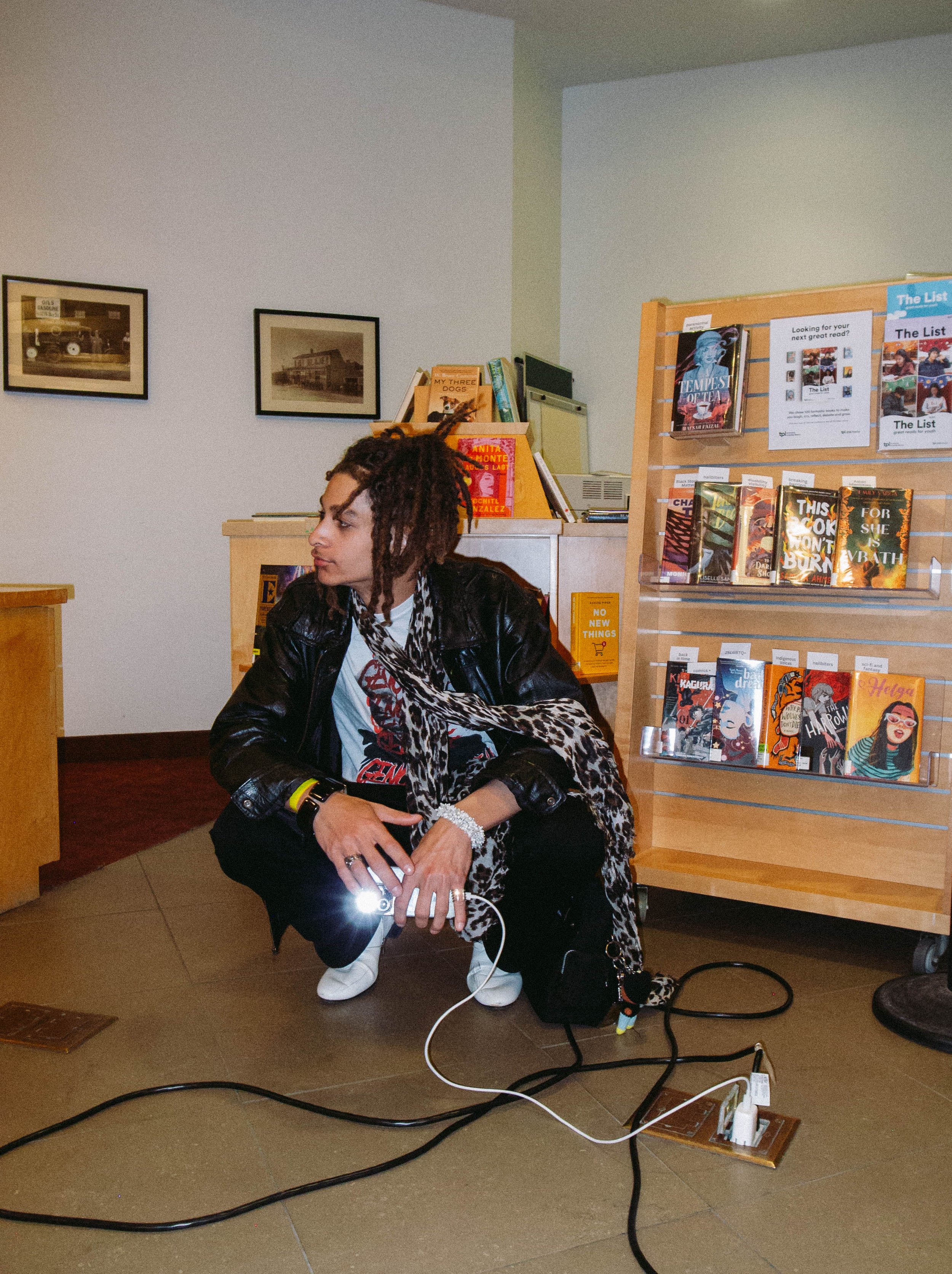 A woman squatting in a bookstore or library, holding a phone with a bright light on. She has dreadlocks, is wearing a black leather jacket, a white shirt, and a leopard print scarf. Behind her is a wooden book display with various books, and framed p