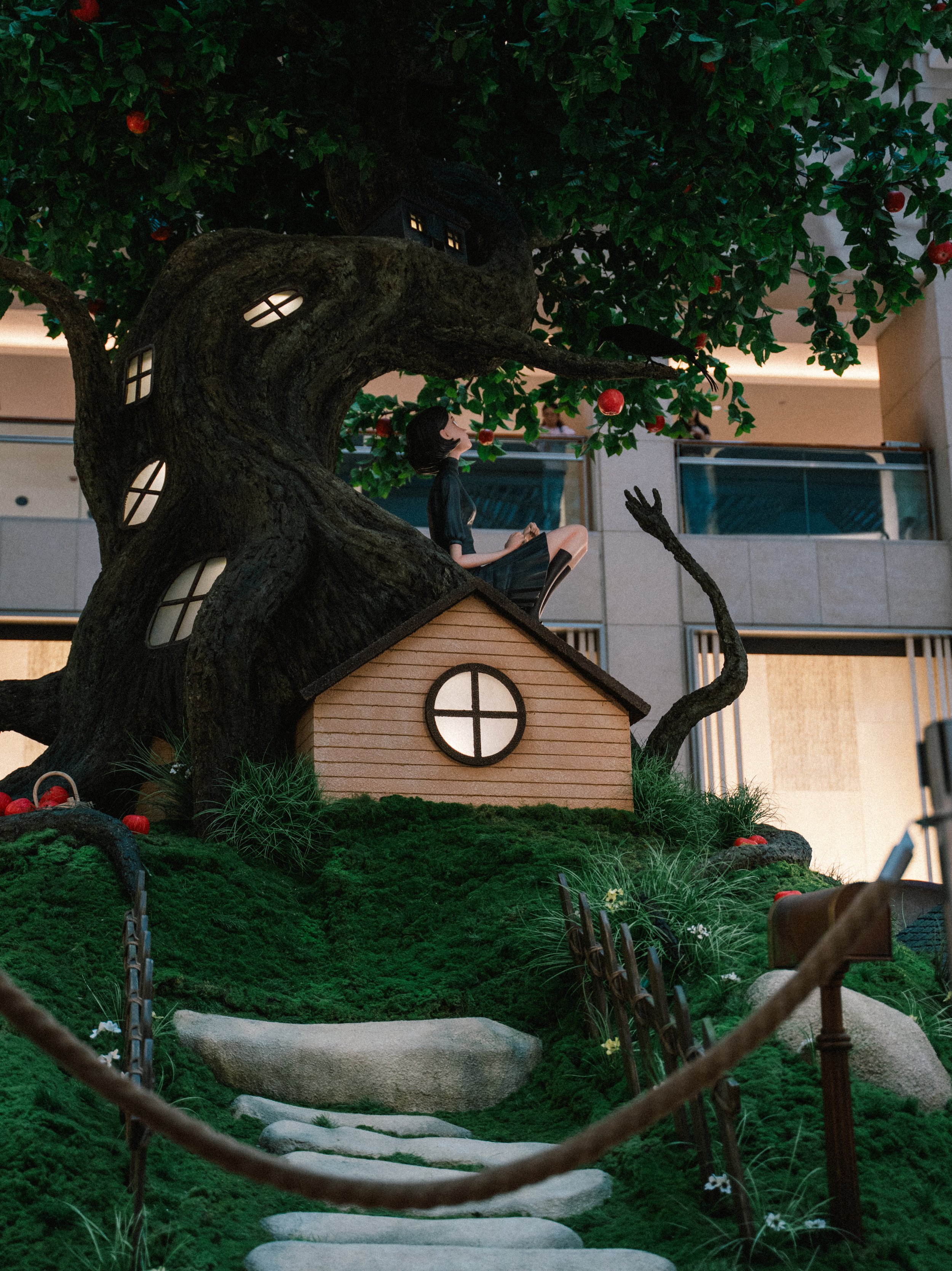 A decorative scene featuring a large artificial tree with windows, a small house at its base, and a girl sitting on the roof. The tree has red apples and greenery, and the scene is set indoors with a modern building background.