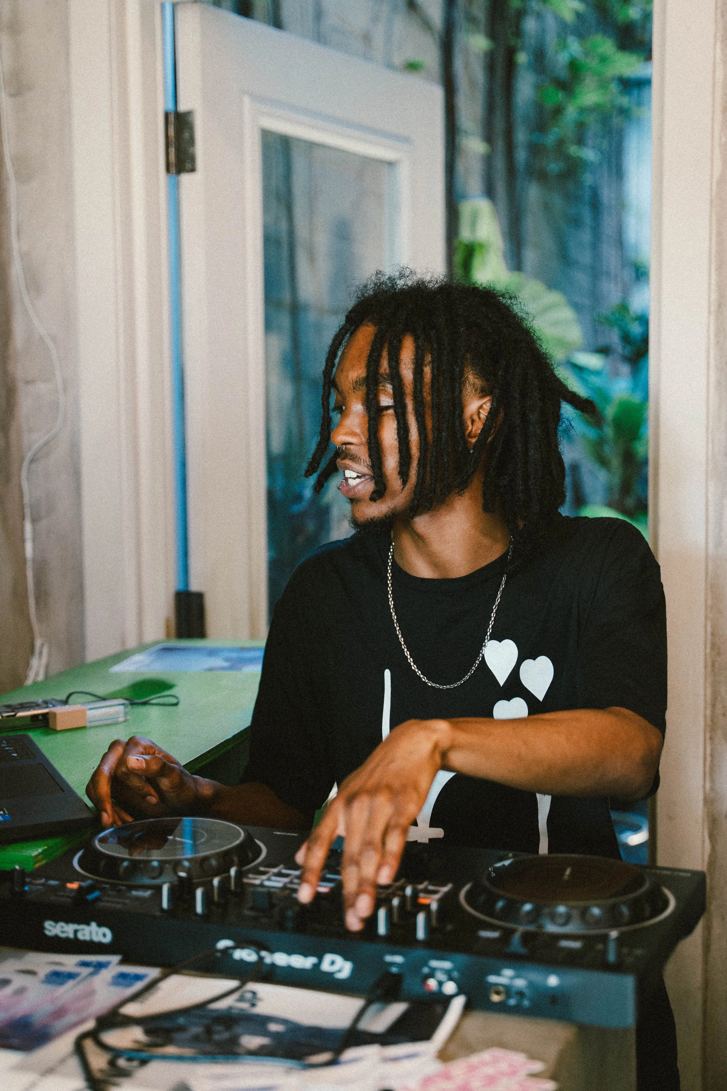 A man with dreadlocks playing DJ at a party, wearing a black shirt with white hearts and a silver chain, inside a room with a green table and a glass door overlooking greenery outside.