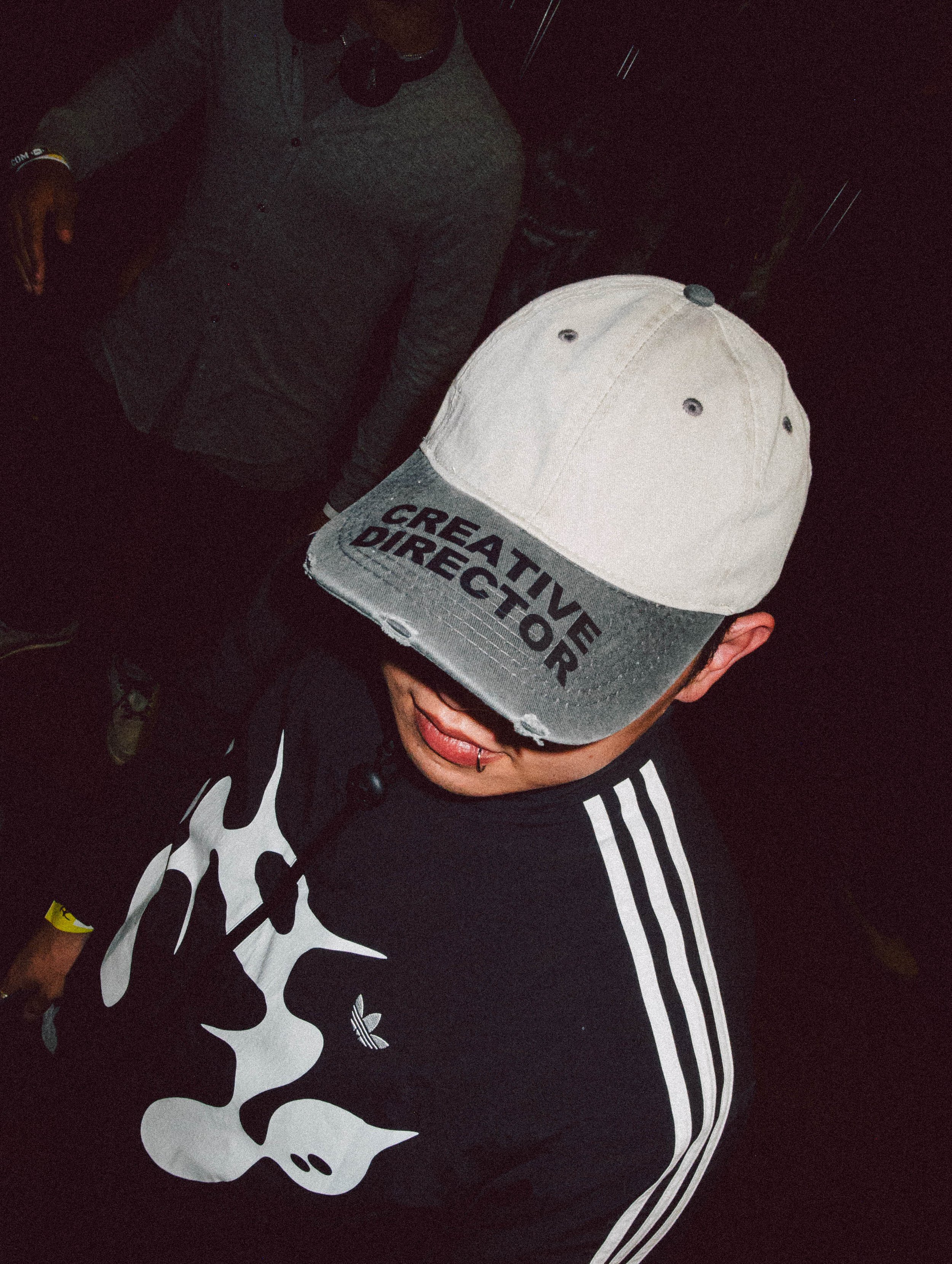 Person wearing a gray and black cap with 'CREATIVE DIRECTOR' written on the front, a black shirt with a white graphic design, and a tongue piercing. The person is looking downward, with a dark background and another individual partially visible in th