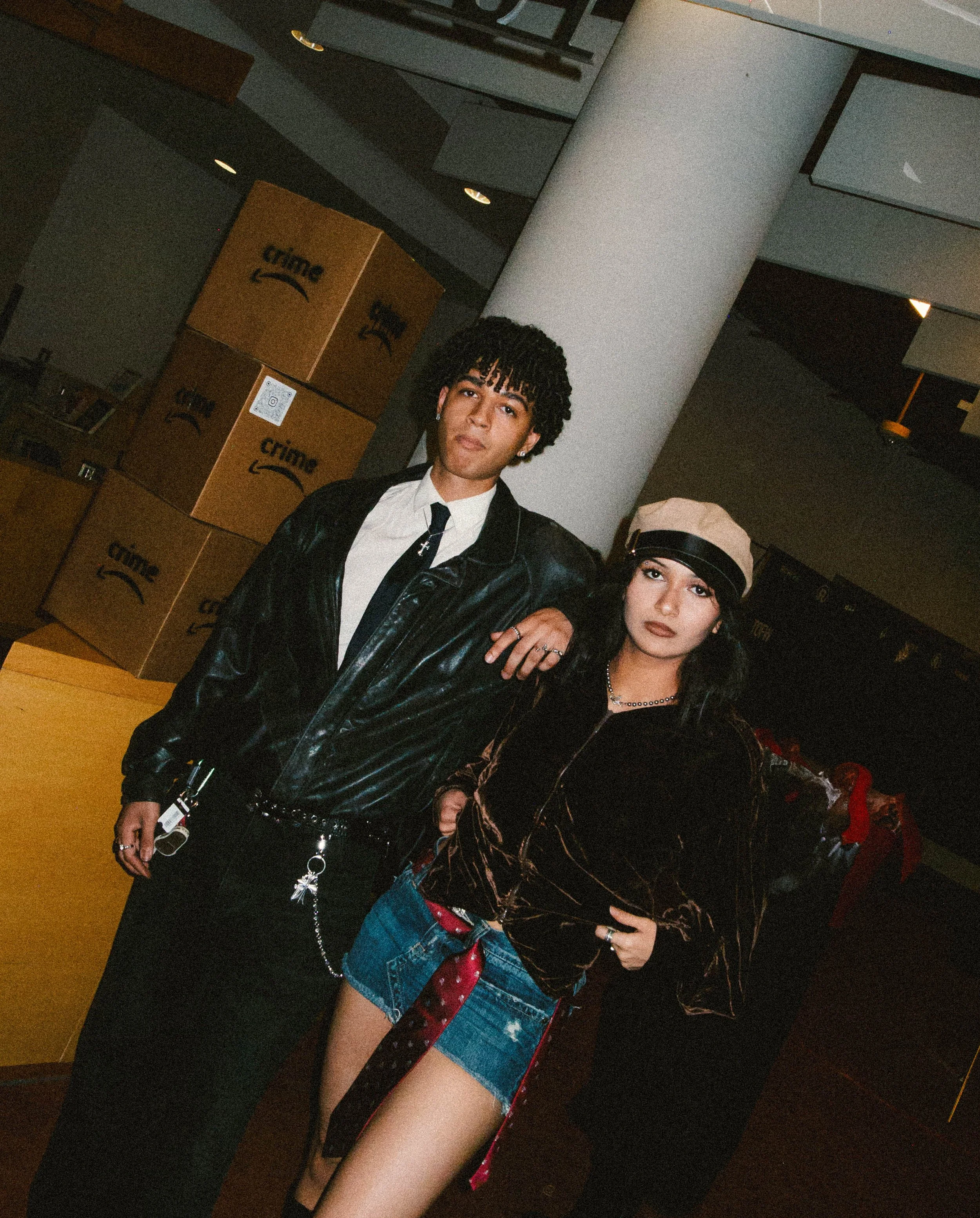 Two young adults, a man and a woman, standing inside a retail store with Amazon boxes in the background. The man has curly hair, is dressed in a black leather jacket, white shirt, black tie, and black pants. The woman is wearing a beige cap, a dark v