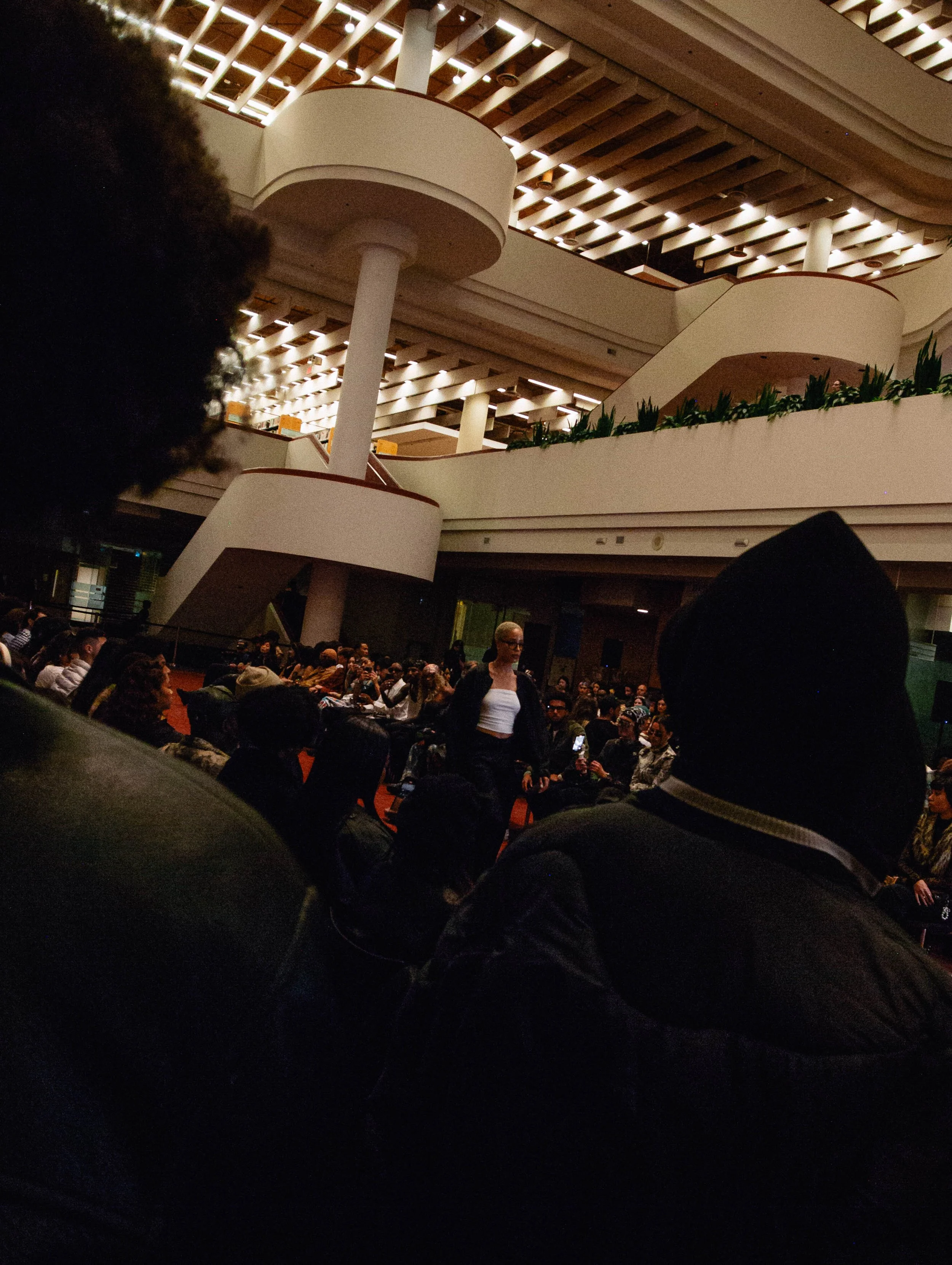 A large indoor gathering with many people seated and standing in a multi-level modern building with architectural balconies and lighting.