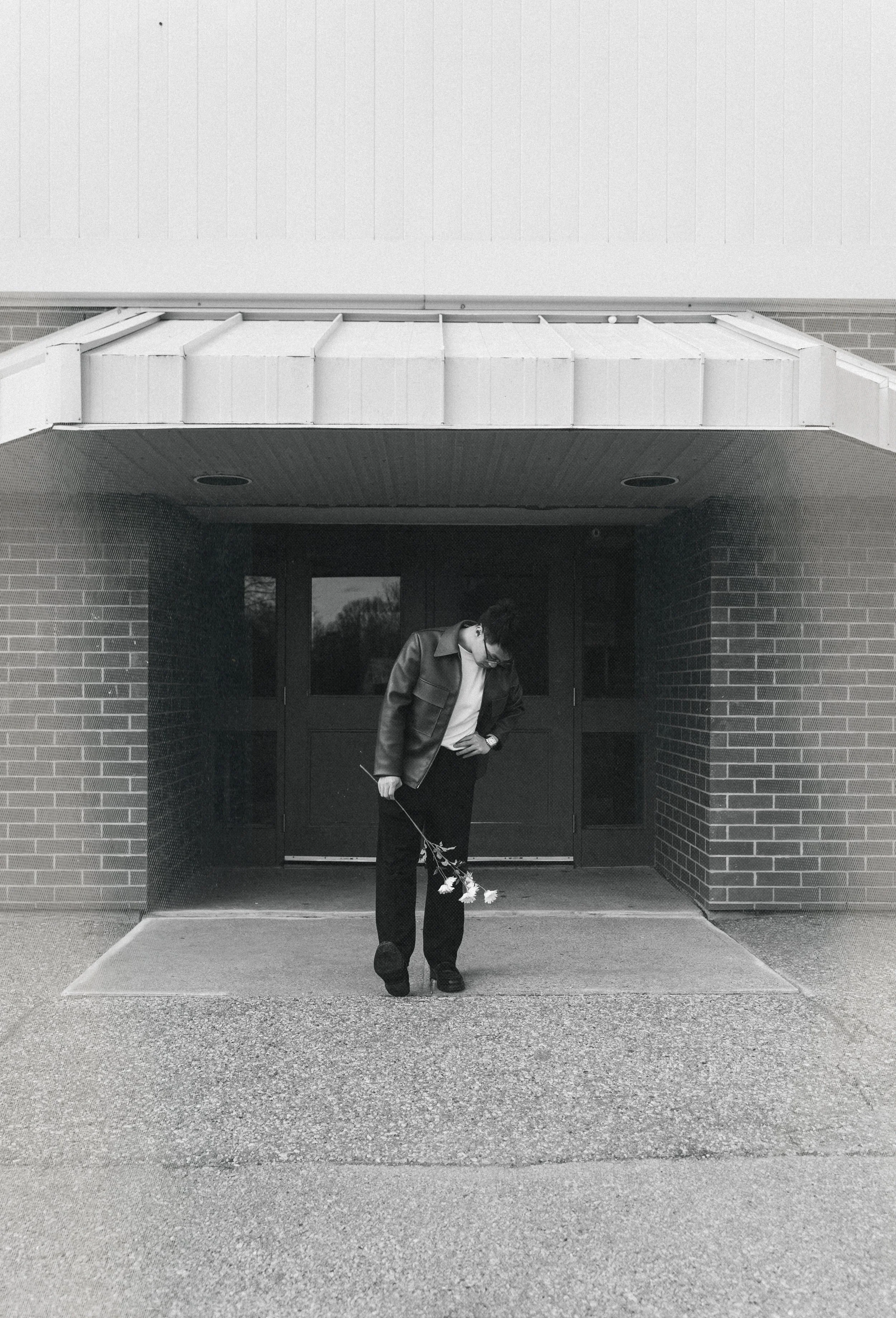 A young man in a leather jacket stands under a building entrance, holding a small bouquet of flowers, looking down at it, in black and white.