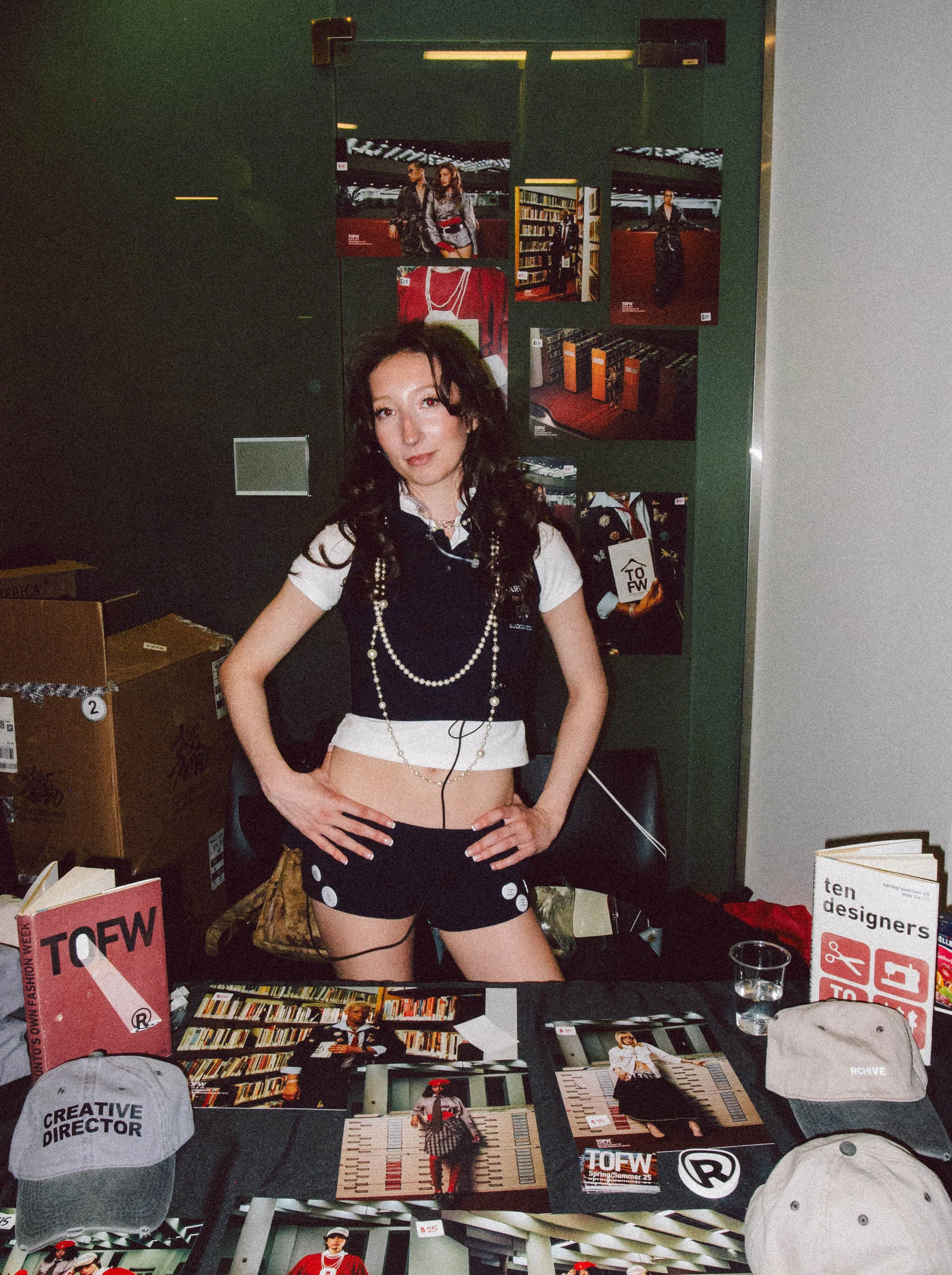 A young woman standing behind a table at a book or fashion event, with photos, books, and caps displayed on the table. She has dark wavy hair, wears a black and white outfit with layered necklaces, and poses with hands on hips.