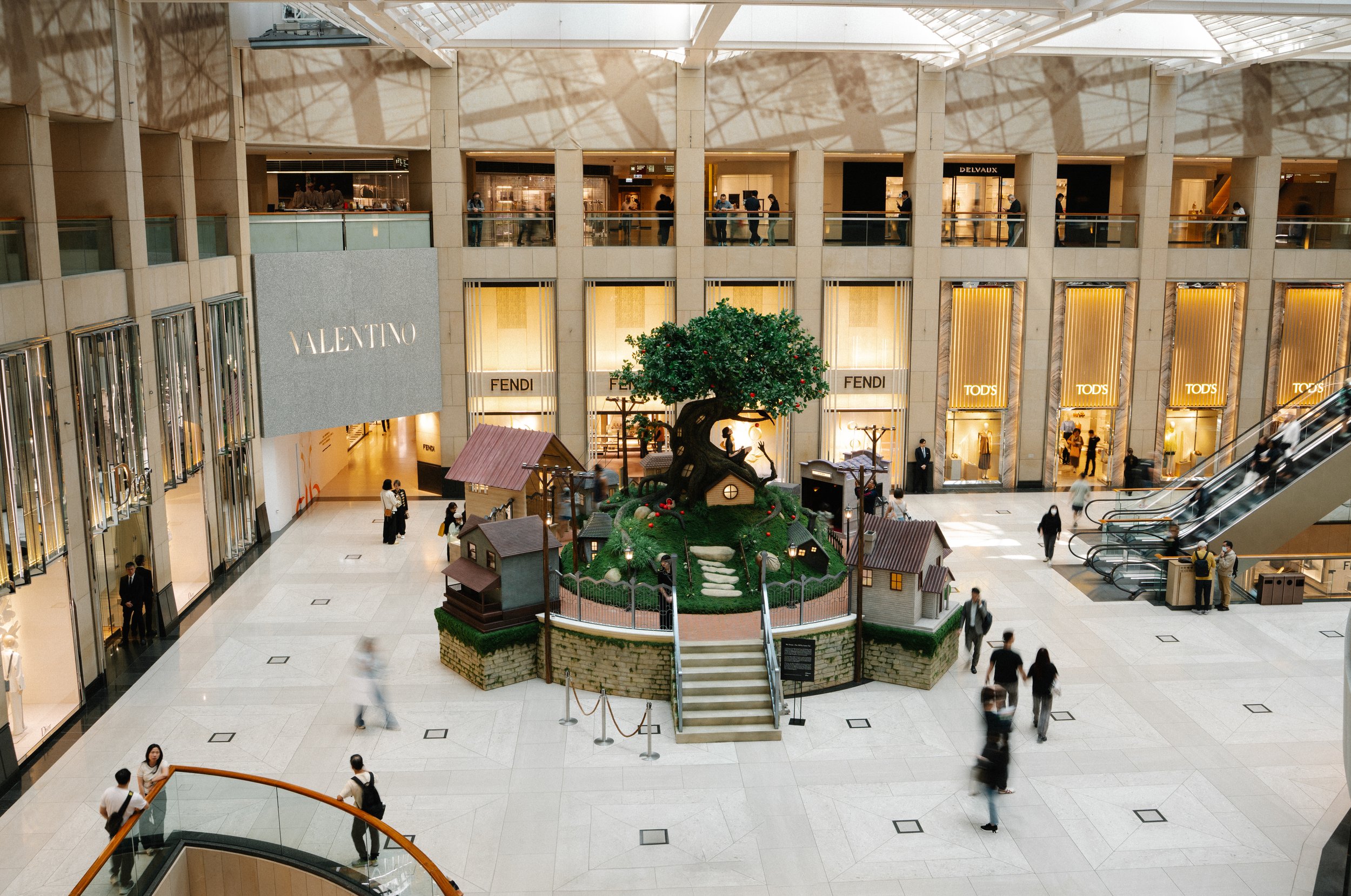 Interior view of a shopping mall with luxury brand stores like Valentino, Fendi, and Tod's. There is a central decorative display with a large tree, small houses, and figurines. People are walking around, some on escalators and some on the ground lev