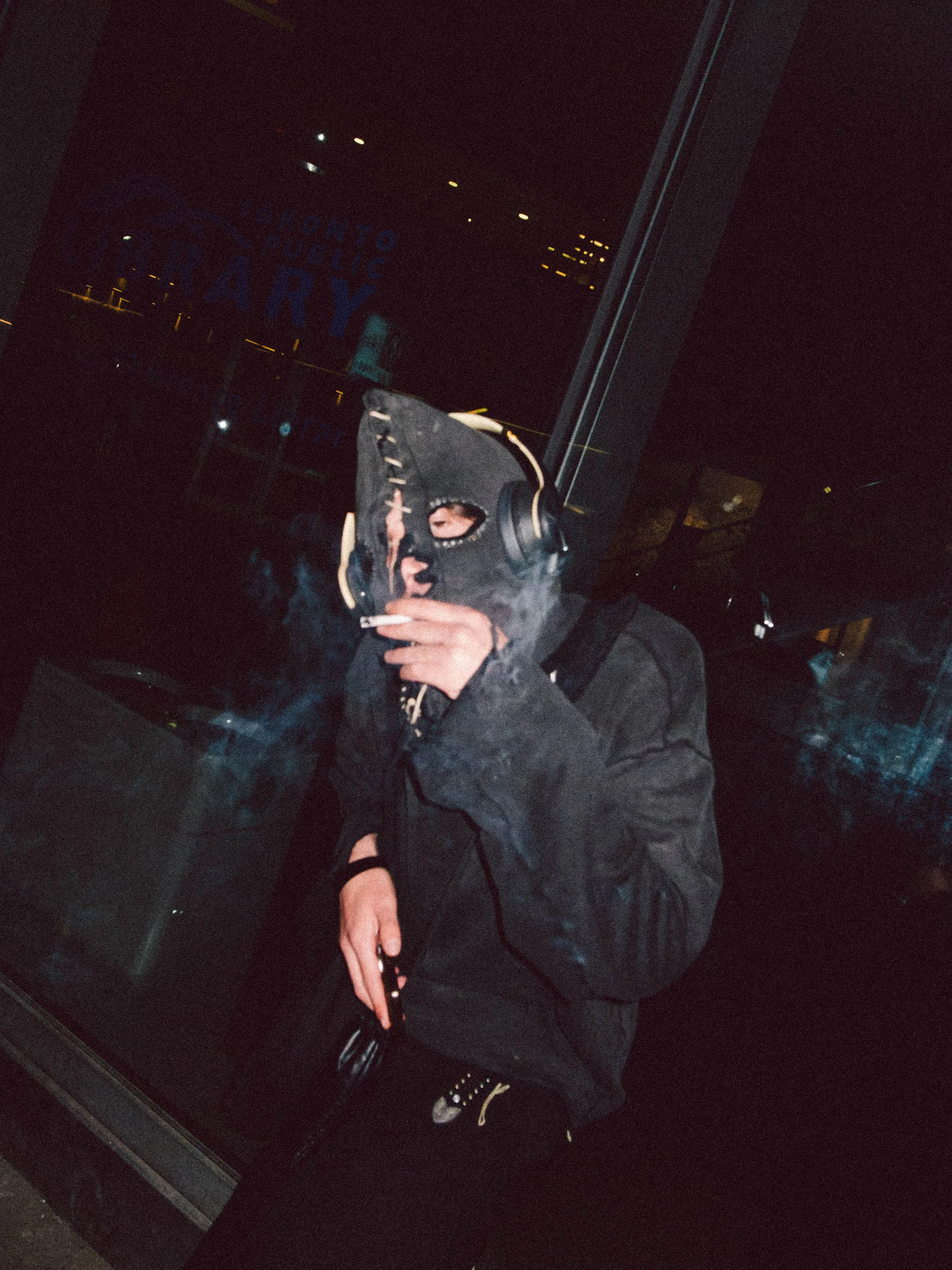 Person wearing a ski mask smoking and holding a cigarette, in a dark environment with city lights reflected on a glass window behind.