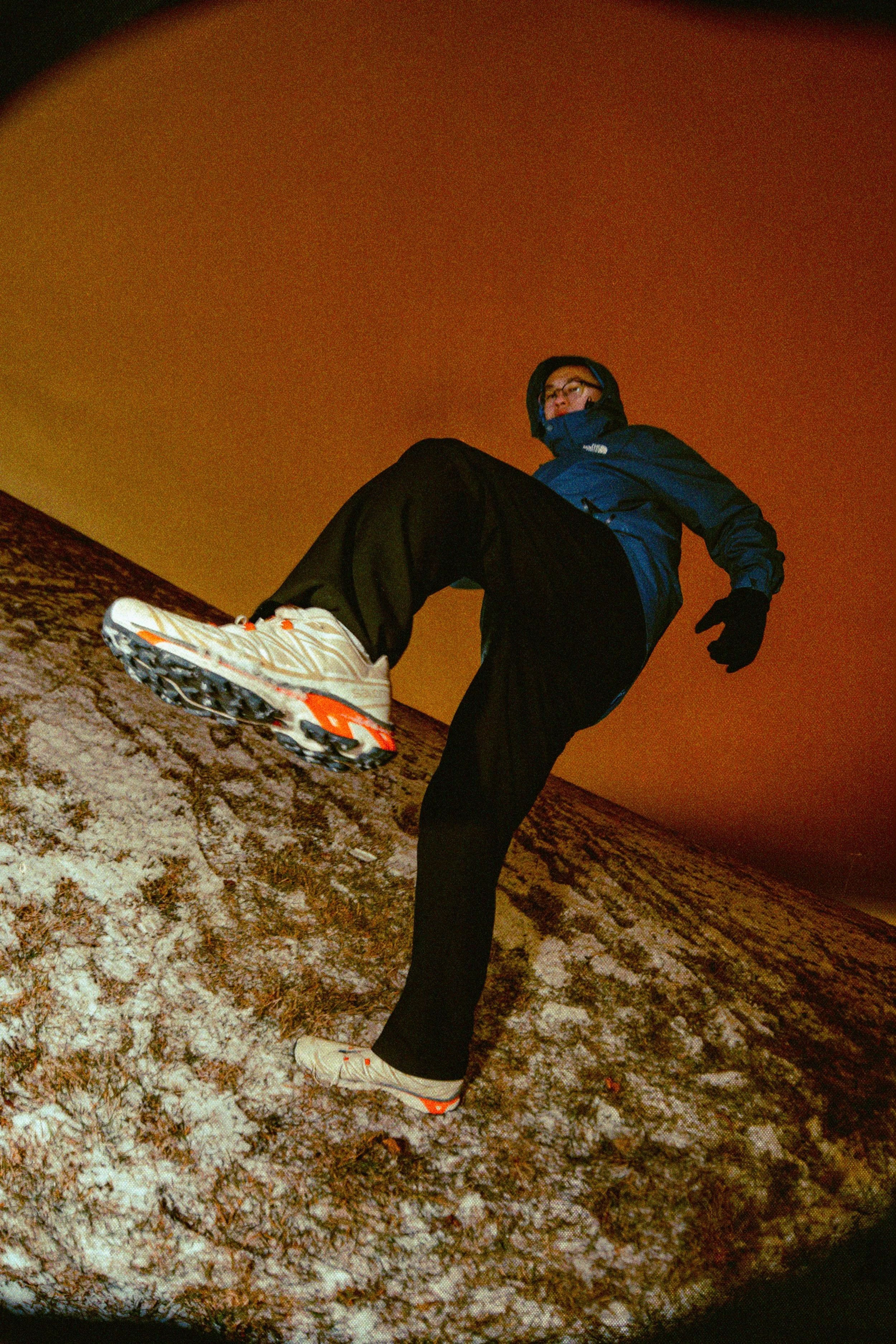 Person in sporty winter clothing jumping or posing on a snow-covered hill at night with an orange sky background.