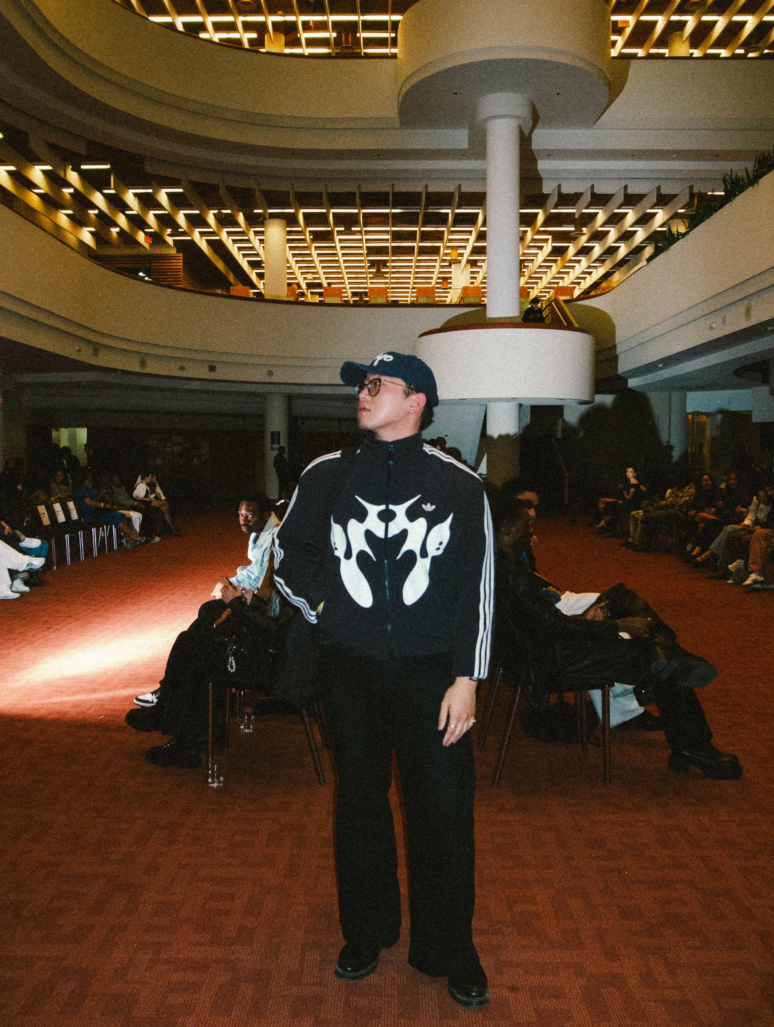 A woman in a black and white jacket with a bold graphic design, black pants, and a baseball cap standing in a large, modern indoor space with seating and multiple floors.