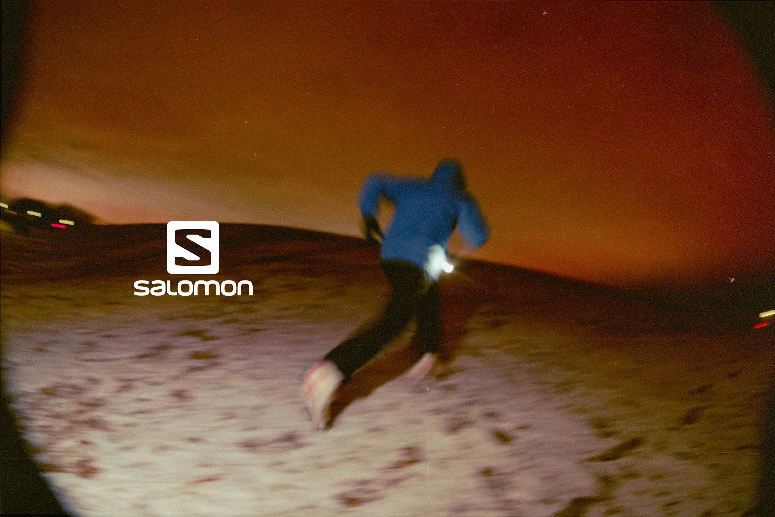 A person in a blue jacket running or jogging on a snowy trail during twilight or early evening. The background has a dark sky with some distant lights and a reddish hue near the horizon. There is a white 'Salomon' logo in the foreground.