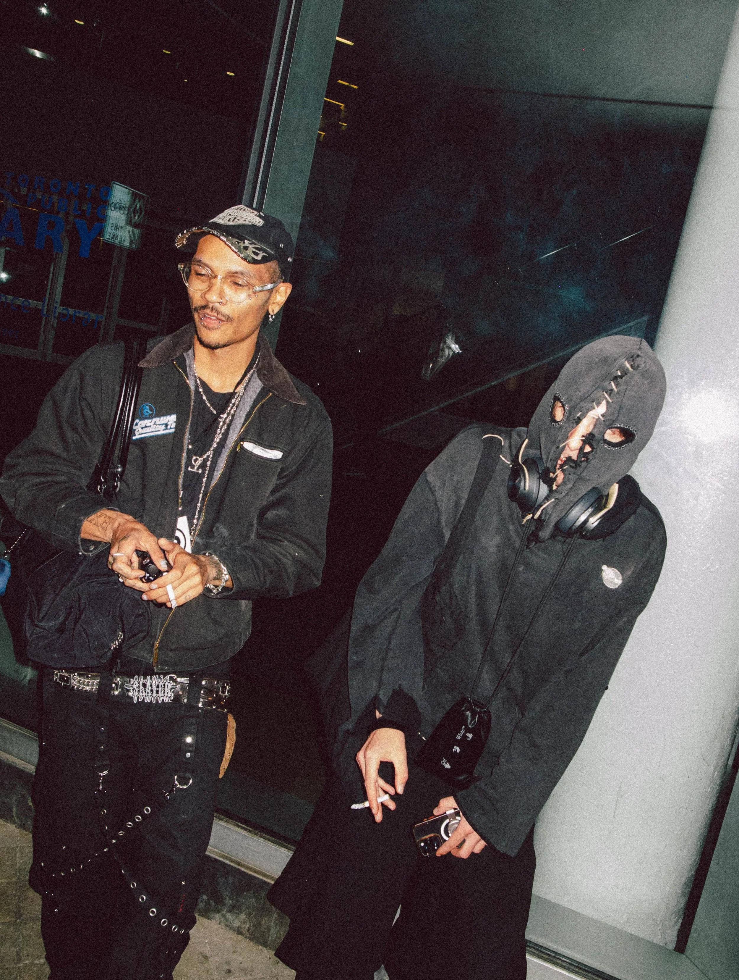 Two young men standing outside at night, one with a cap and jewelry, the other wearing a ski mask with holes and headphones around his neck, both holding cigarettes.