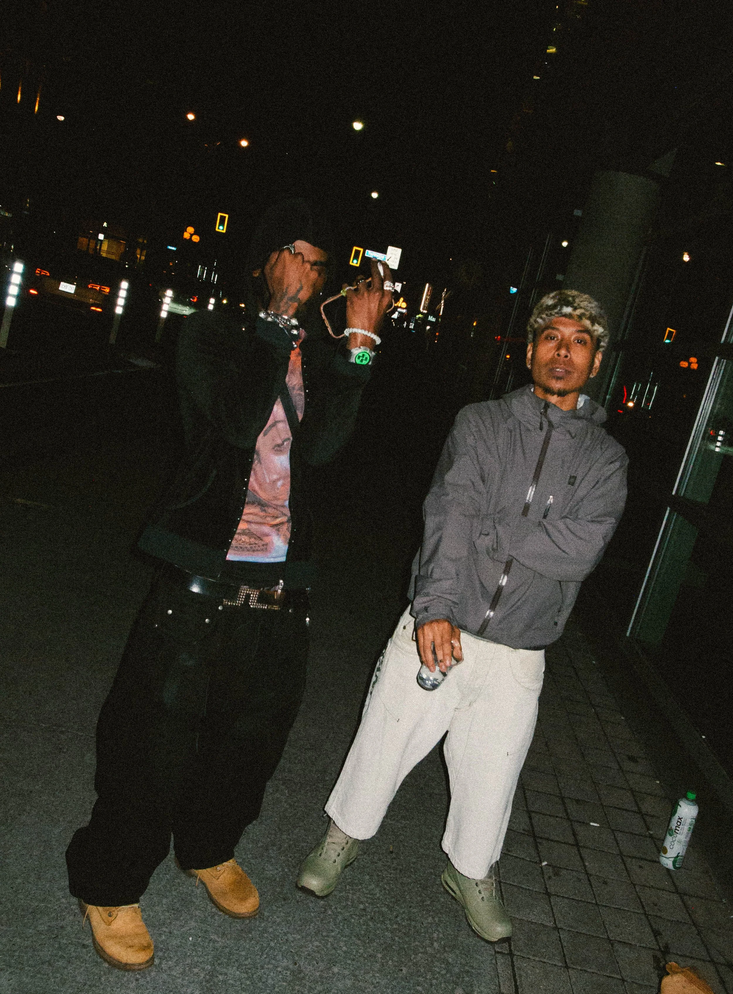 Two young men standing on a city street at night, with one taking a selfie photo. They are dressed casually with jackets, pants, and boots, and are illuminated by streetlights and city neon signs.