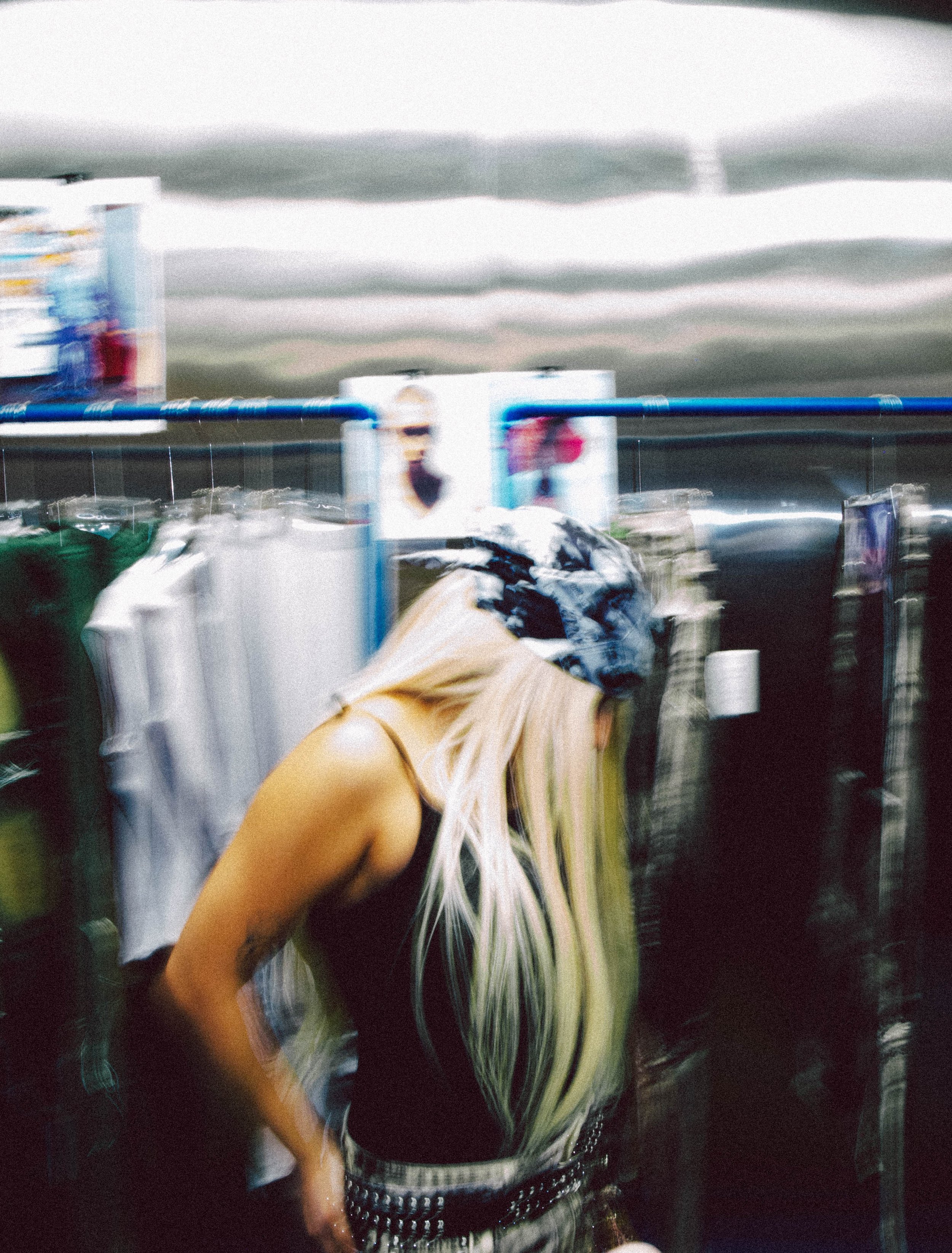 A woman with platinum blonde hair wearing a black tank top, patterned belt, and a colorful bandana on her head, shopping for clothes in a store with blurred clothing racks and advertisements in the background.  the texture is also filmic 