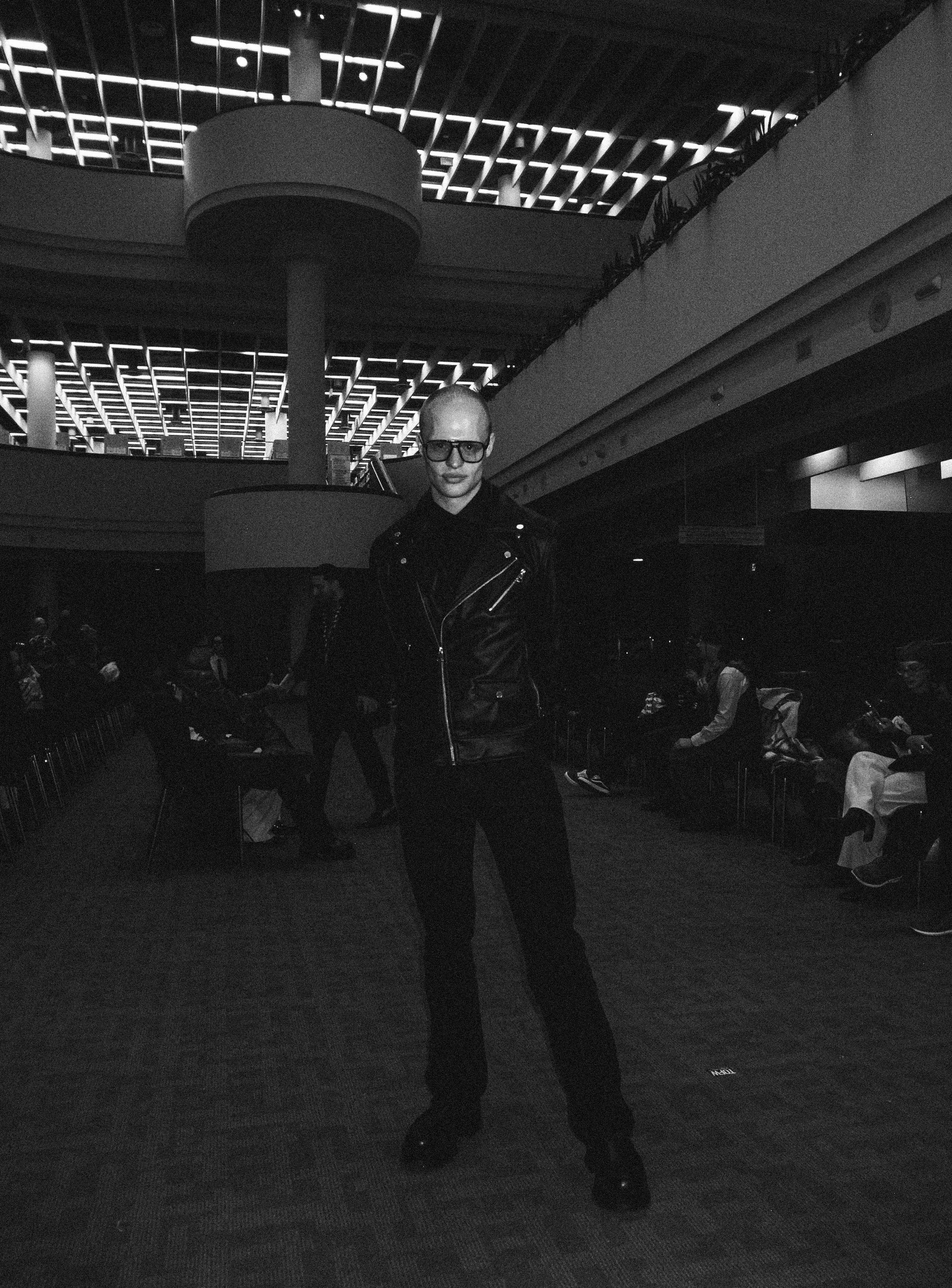 A man with light skin, bald head, sunglasses, wearing a black leather jacket and black pants, standing in an indoor public space, possibly an airport or convention center, with people sitting in chairs in the background and modern architectural desig